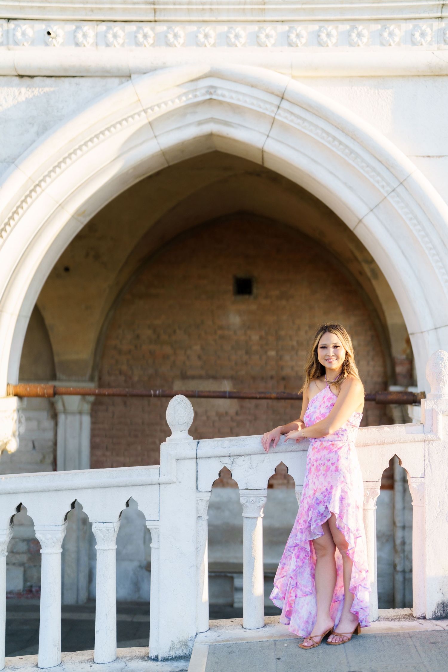 Young woman in pink floral dress posing by white ornate balustrade under Gothic arch of historic building.