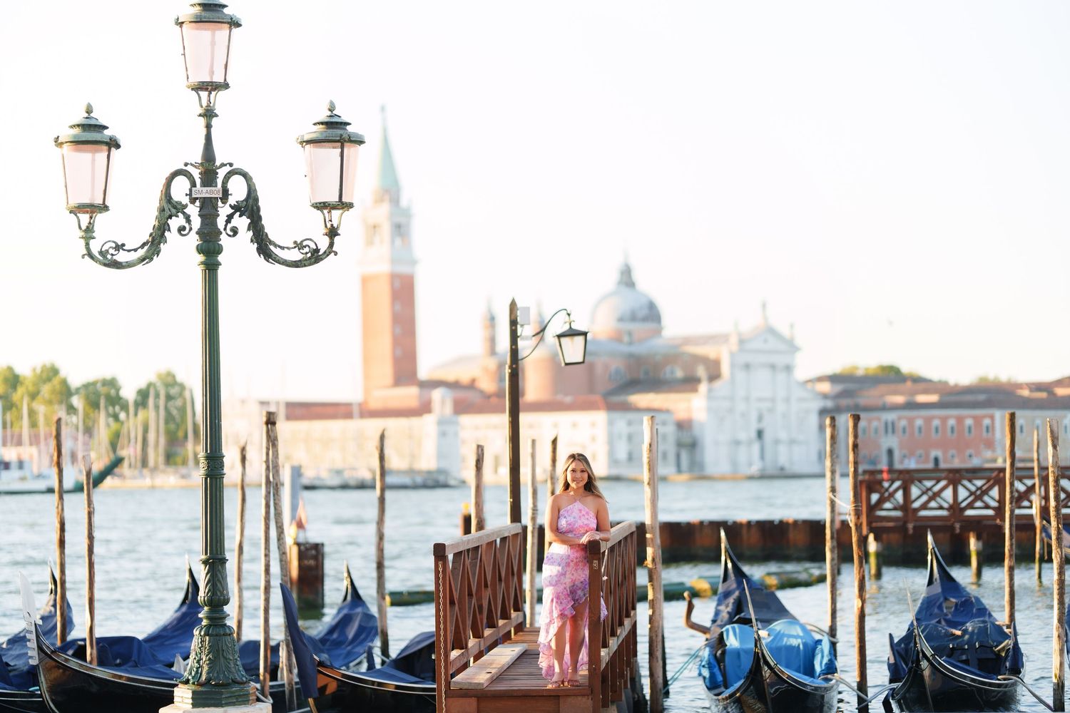 Woman in pink dress standing on Venice gondola dock with San Giorgio Maggiore church and lamp post at sunset.