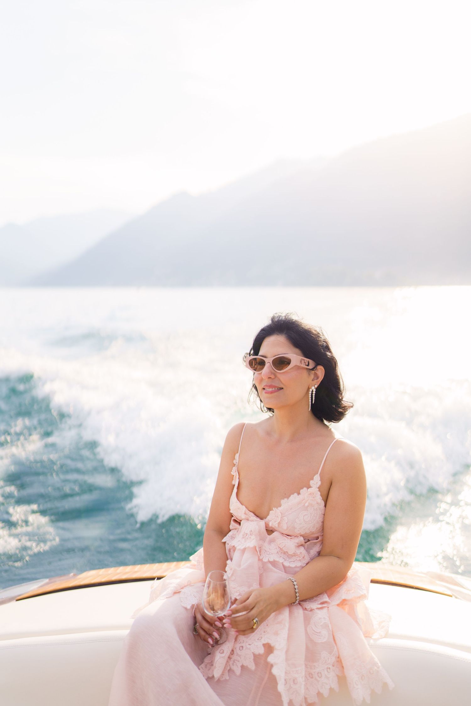 Woman in pink dress sitting on yacht stern with mountain backdrop and ocean wake.