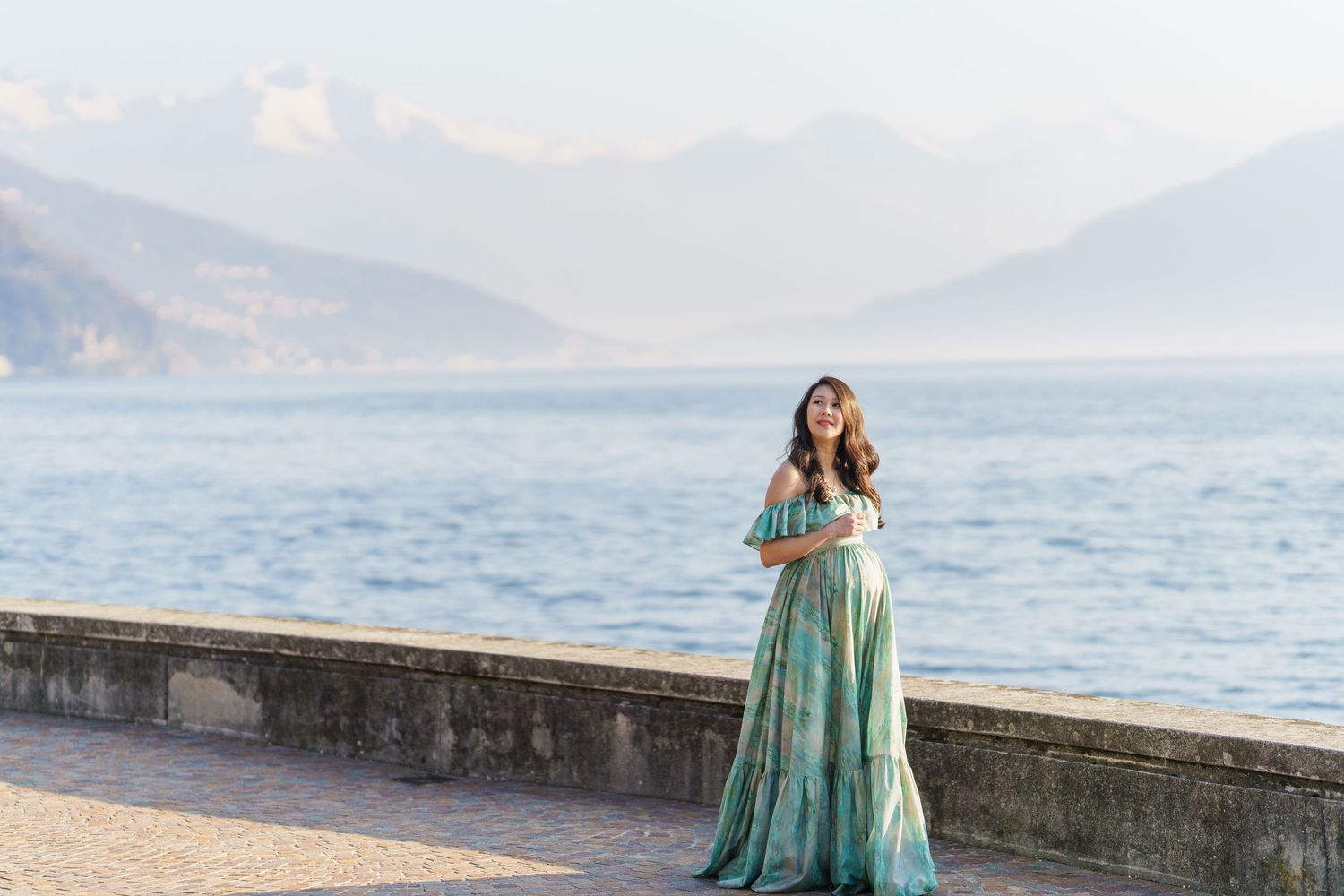 Woman in teal floral maxi dress poses by lakeside promenade with misty mountain backdrop.
