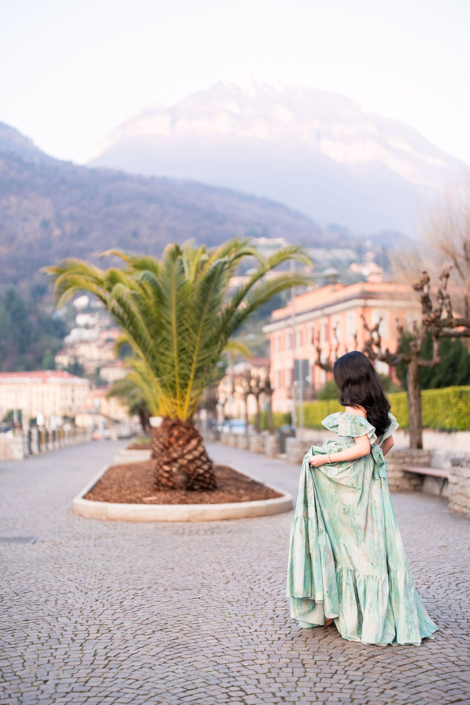 Woman in elegant mint green gown stands on cobblestone promenade with palm tree and mountain backdrop.