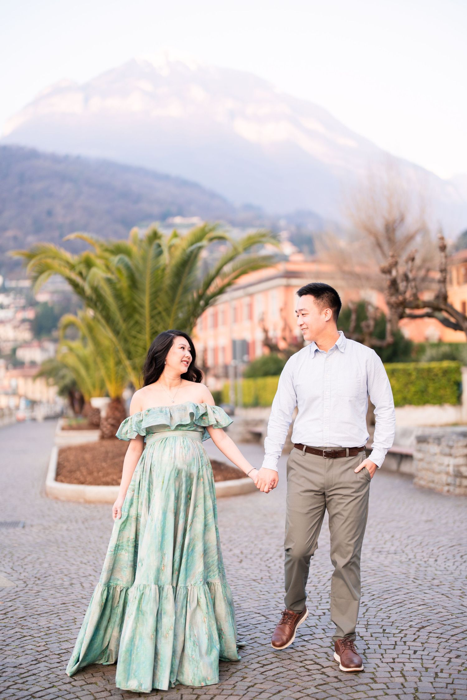 Couple holding hands on cobblestone plaza with palm trees and mountain backdrop, woman in green off-shoulder gown.
