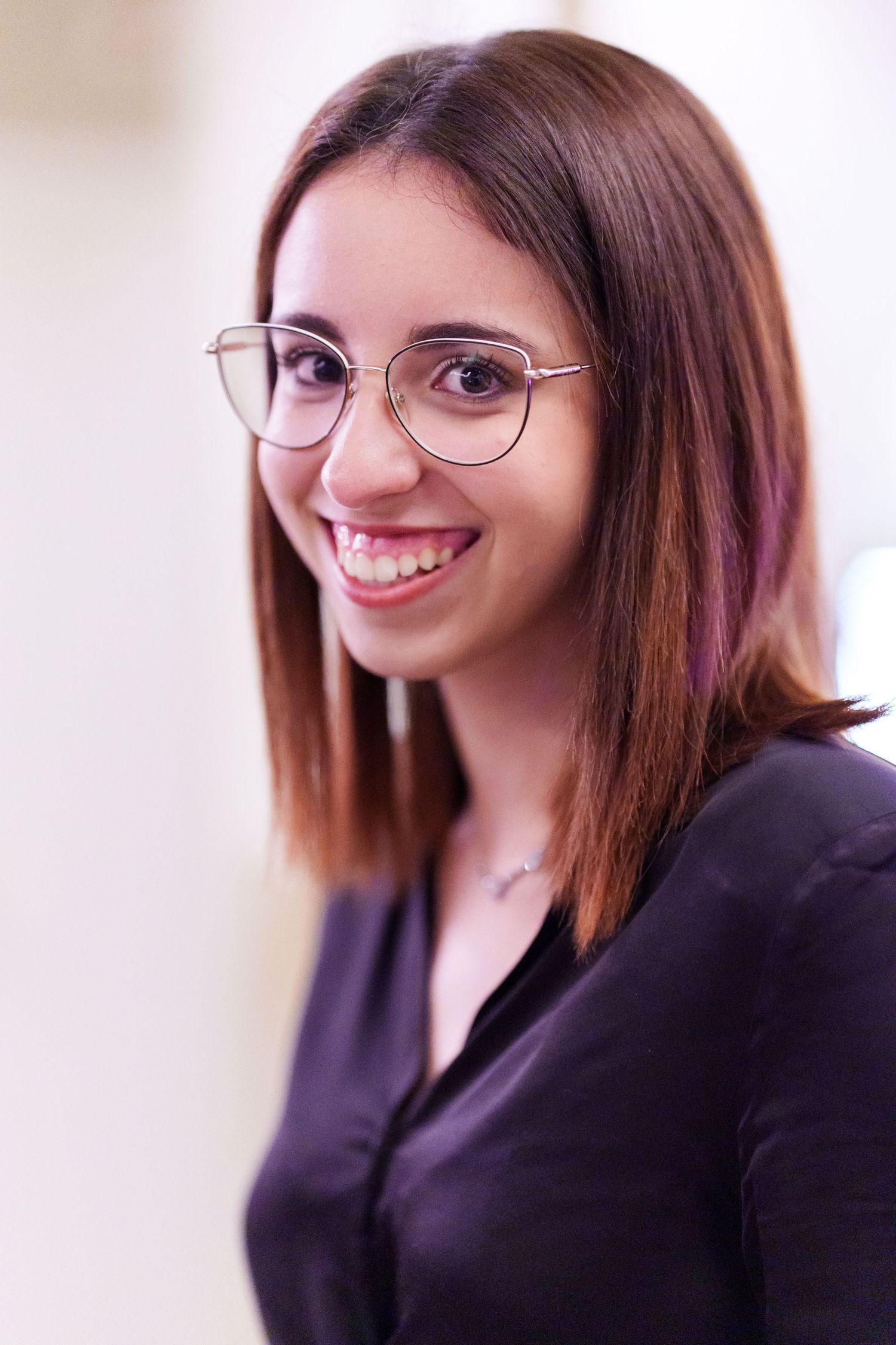 Young woman with glasses and brown hair smiling confidently in a black blouse, professional headshot portrait.