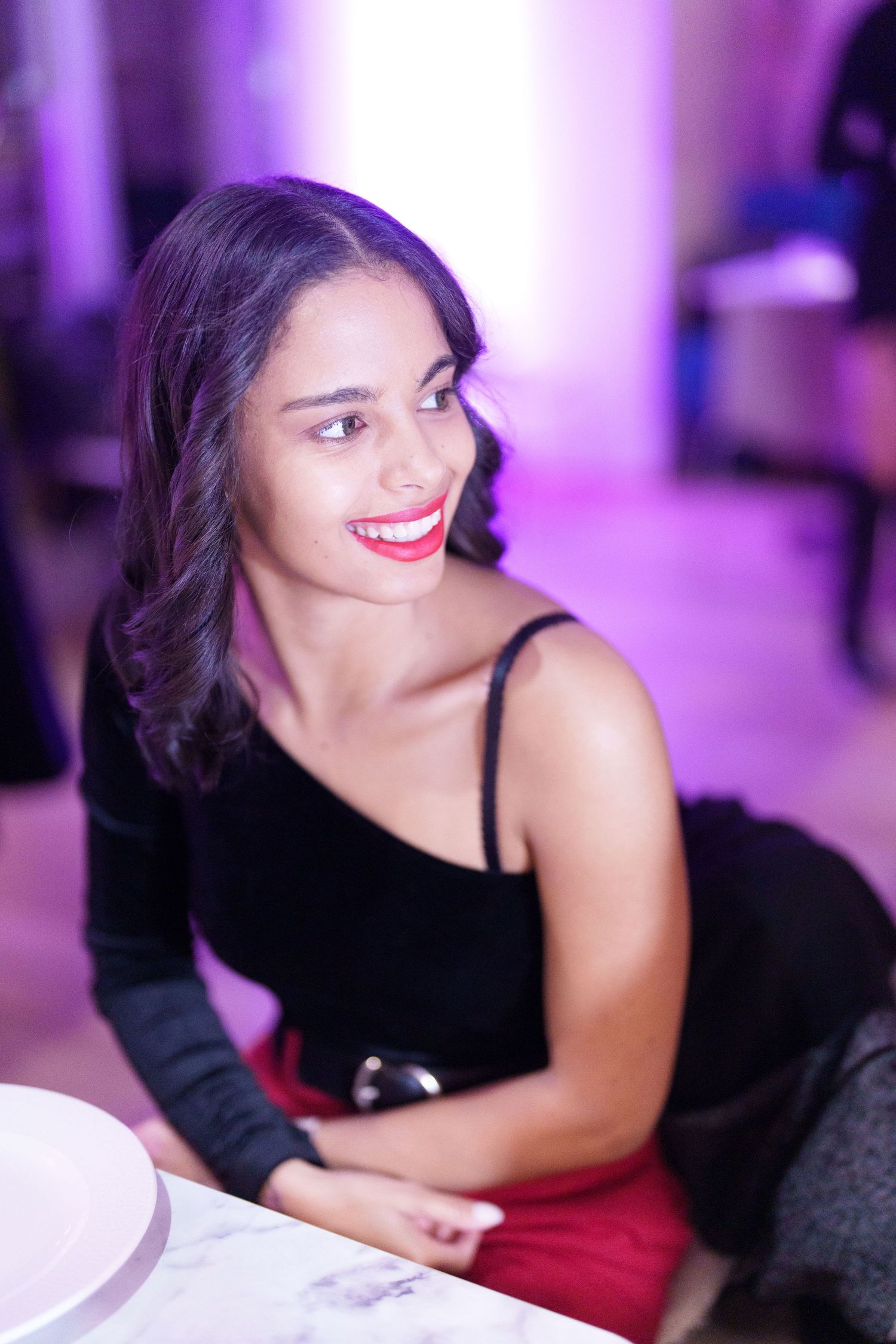 Smiling young woman in black top and red skirt sitting at a table in a purple-lit venue with red lipstick.