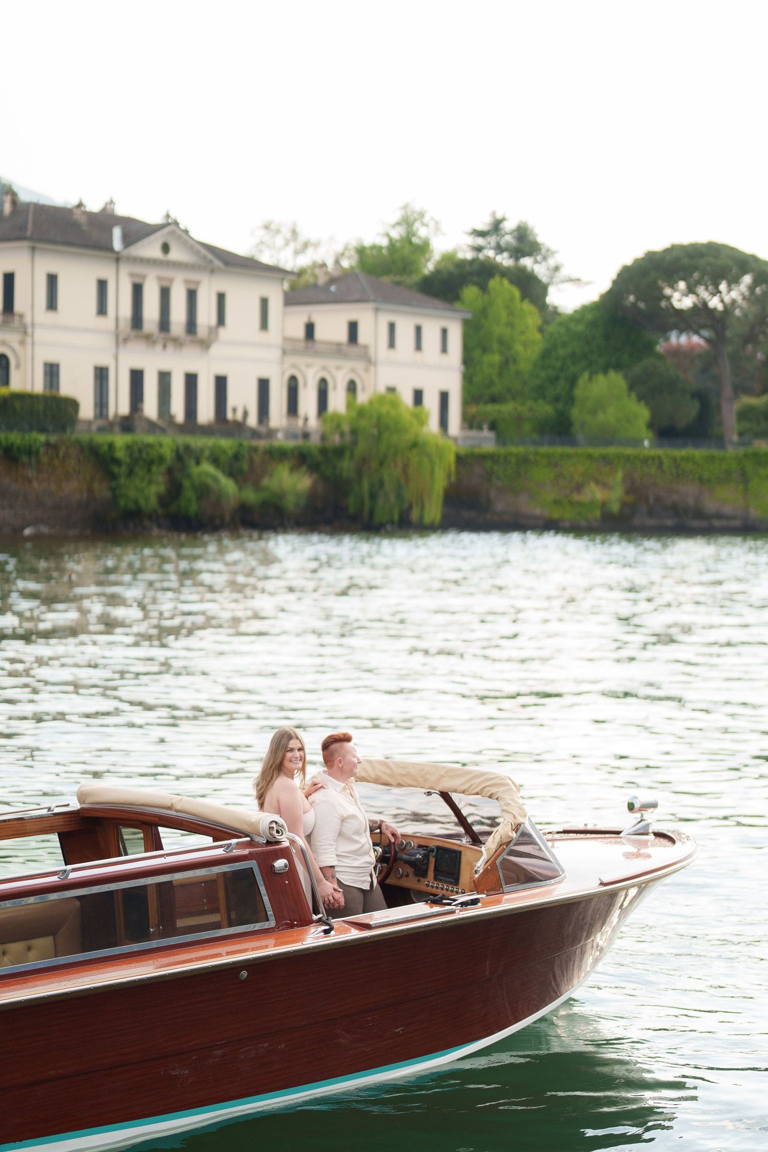 Couple enjoying a romantic boat ride on a wooden vessel with a grand Italian villa and lush greenery in the background.