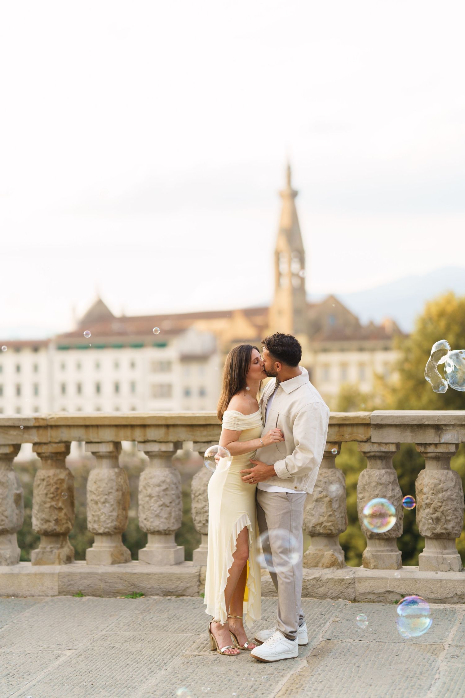 Couple kissing on a stone bridge with soap bubbles floating around them, European cityscape in background.