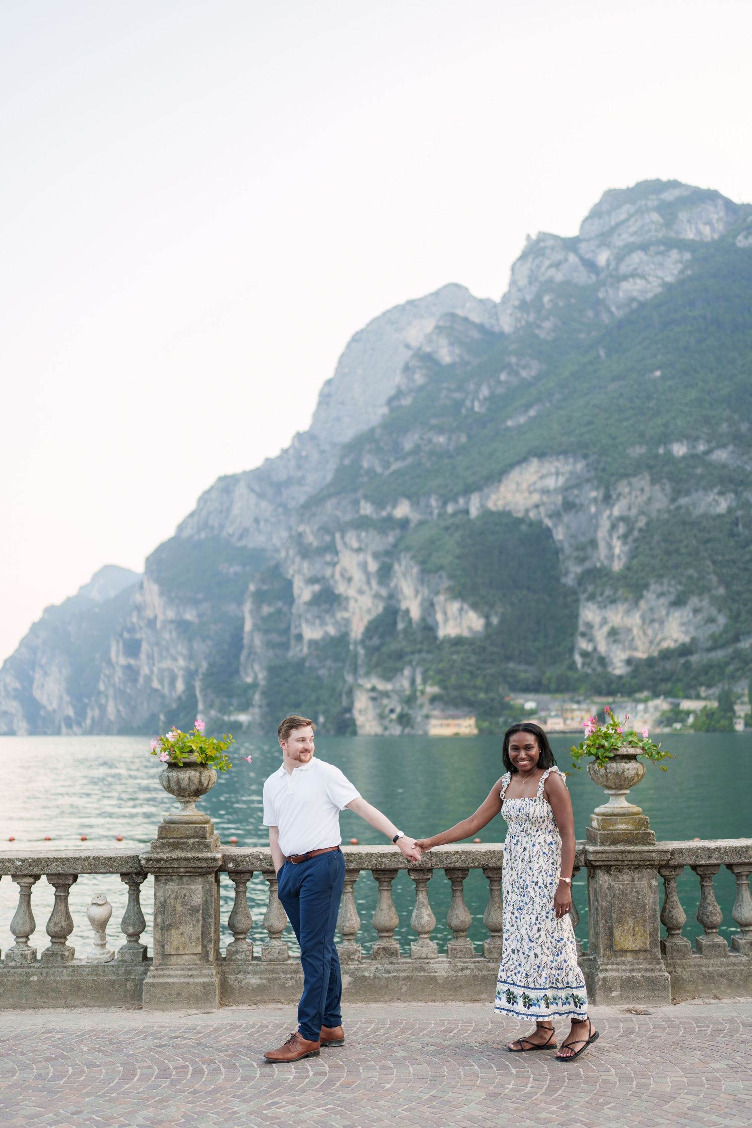 Interracial couple holding hands on a lakeside terrace with dramatic mountain backdrop at Lake Garda, Italy.