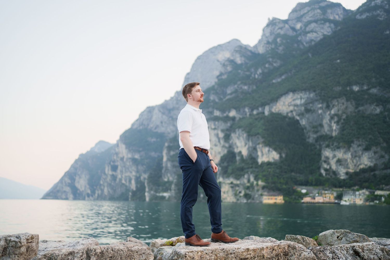 Well-dressed man posing on rocky lakeside with stunning alpine mountains and calm water behind him.