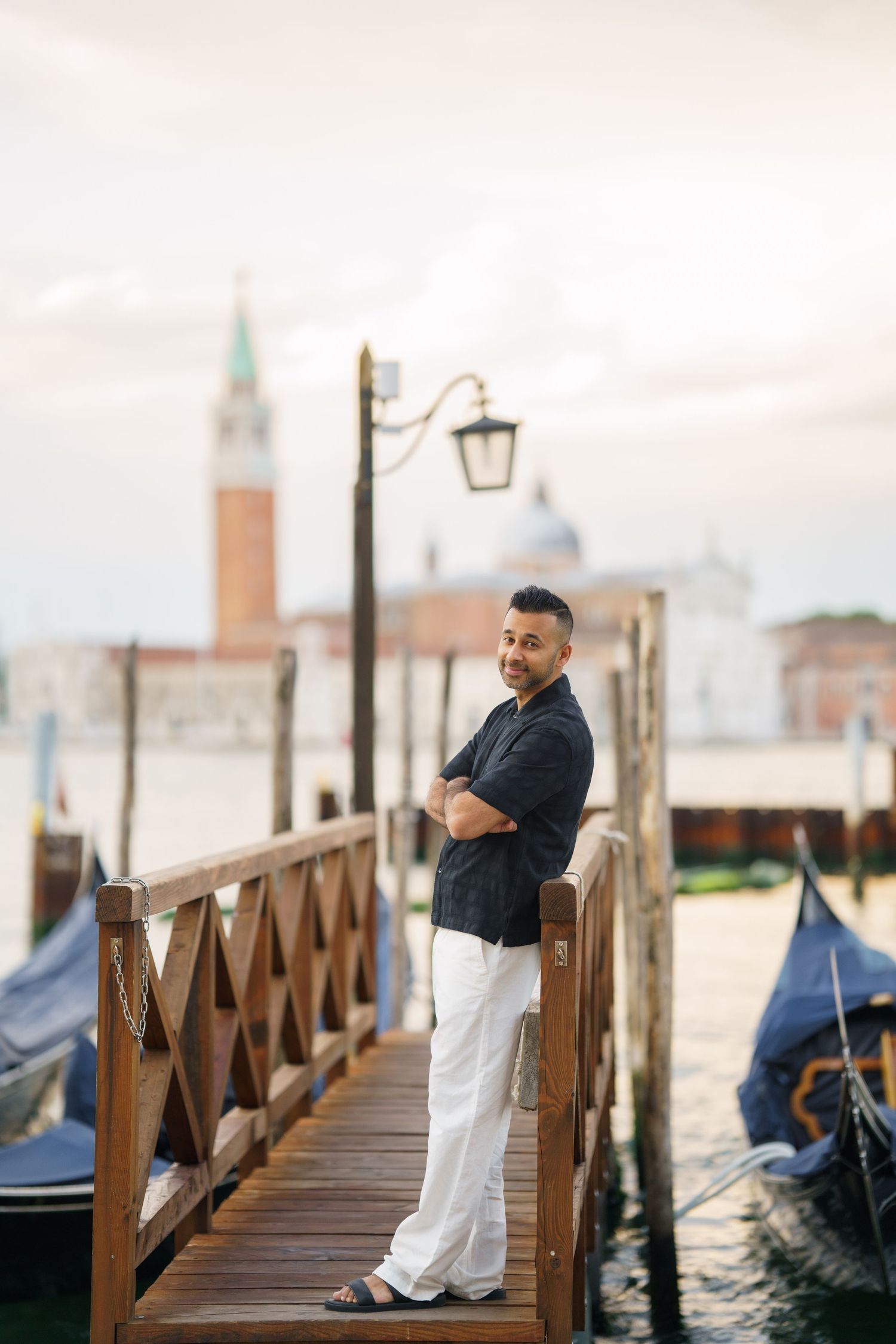 Man in black shirt and white pants stands on Venice dock with San Giorgio Maggiore church in background.