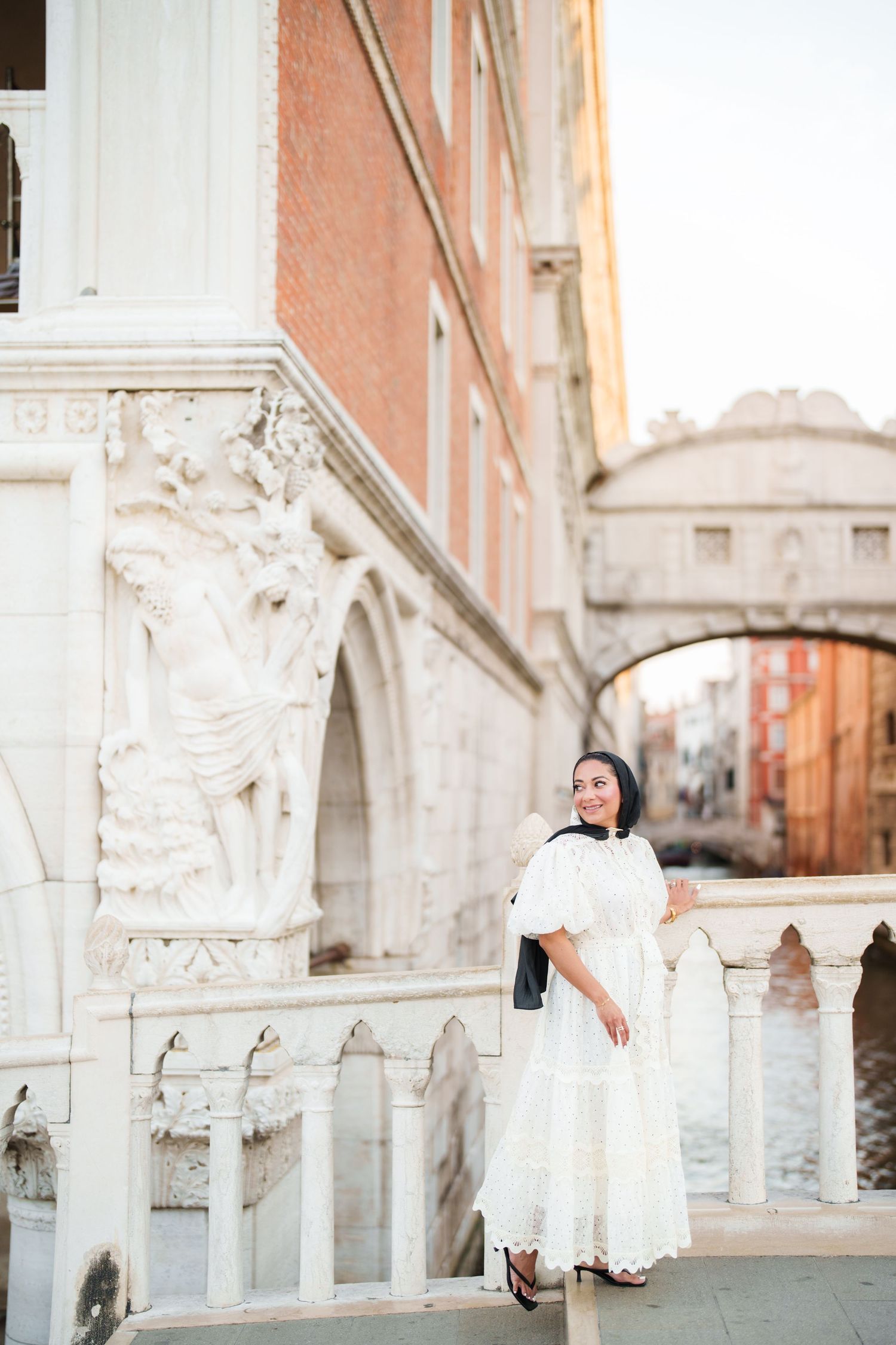 Woman in white dress posing near ornate marble architecture with Bridge of Sighs in Venice, Italy.