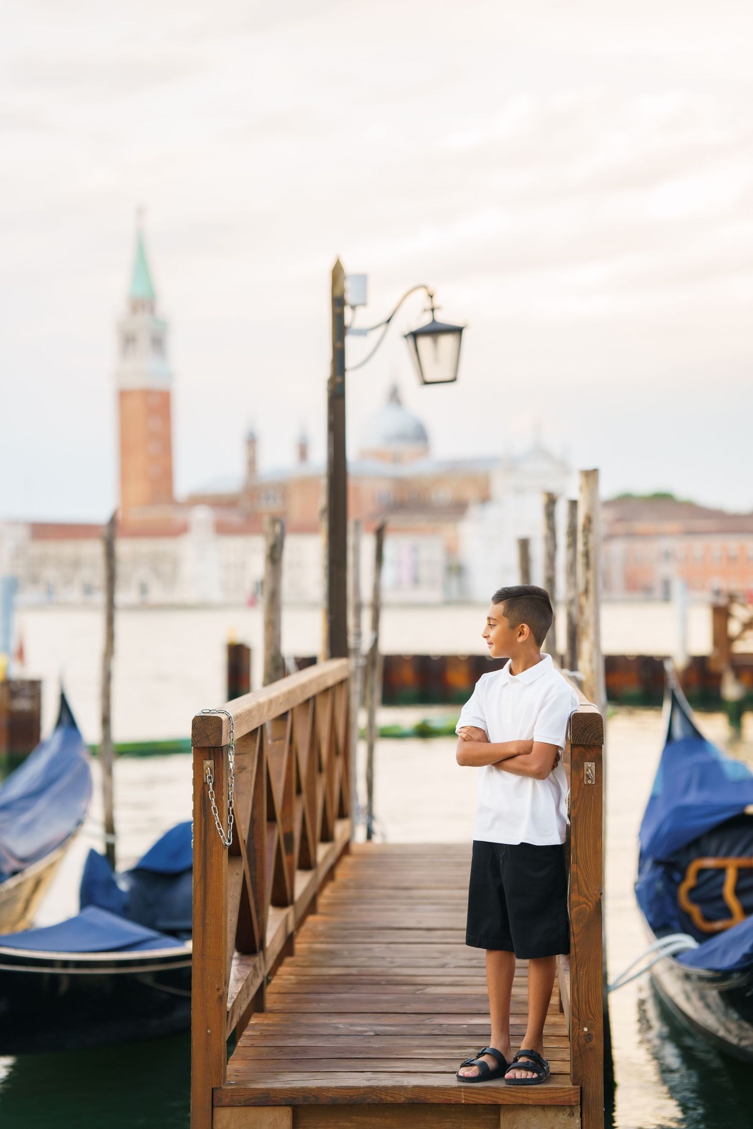 A boy stands on a wooden dock in Venice, Italy, with gondolas and San Giorgio Maggiore island in the background.