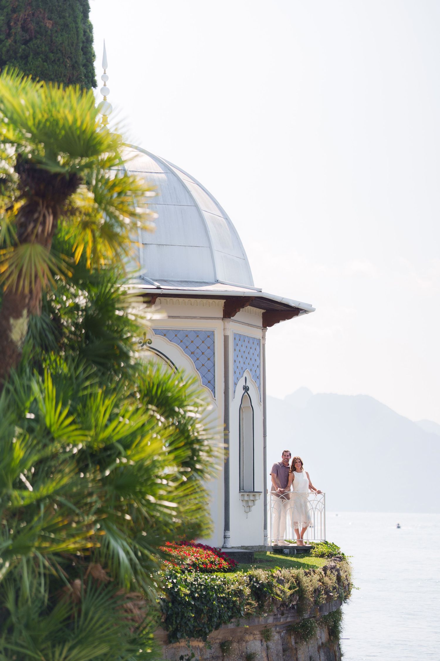 Couple stands on a scenic lakeside pavilion with a domed roof, surrounded by palm trees and mountain views.