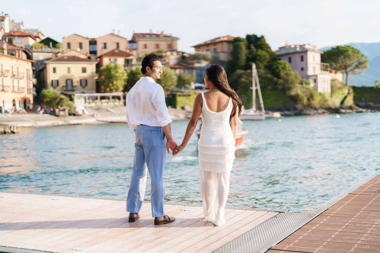 Couple holding hands on a dock overlooking a picturesque Italian lakeside village with colorful buildings.
