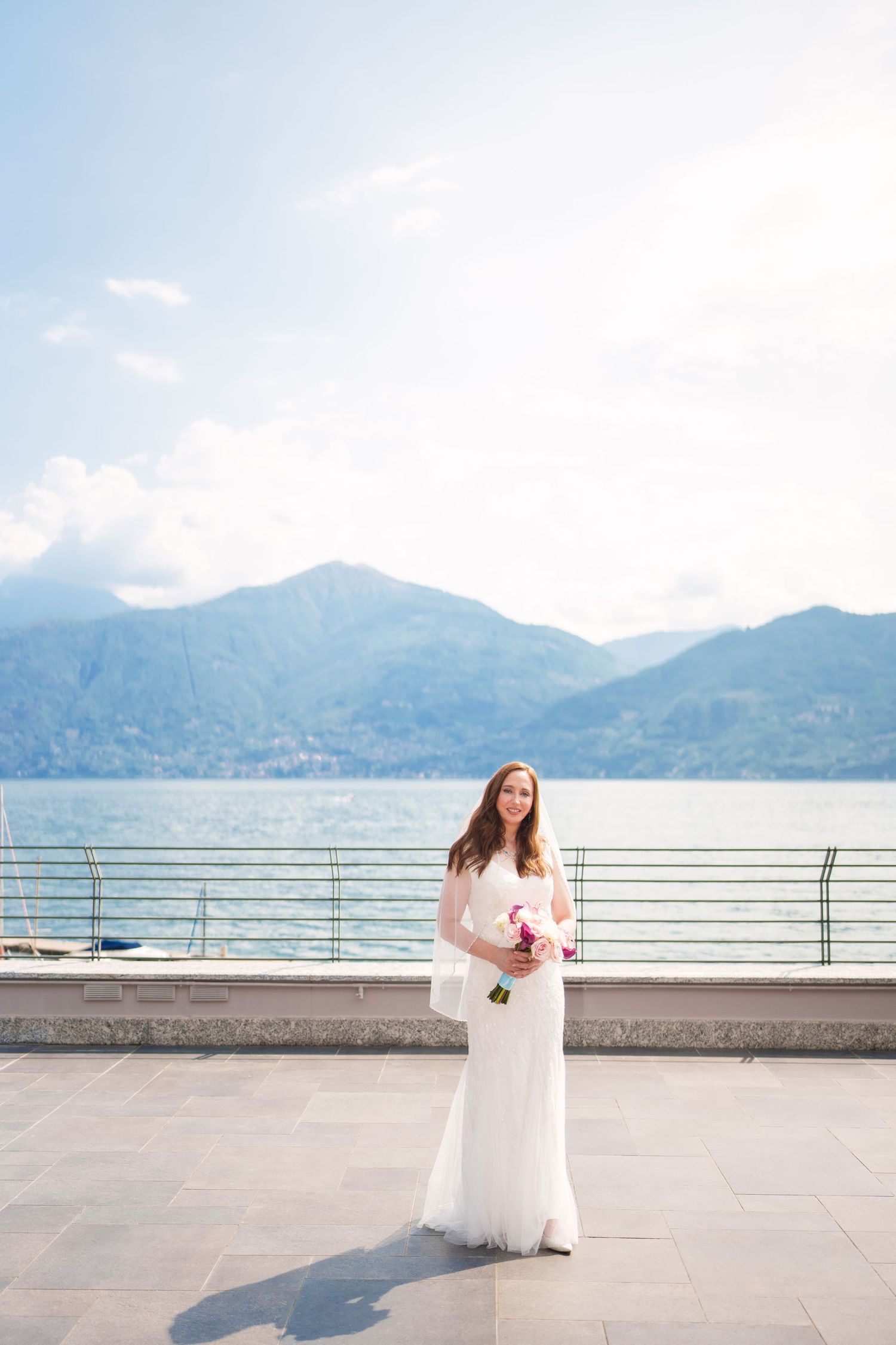 Bride in white gown holding purple bouquet on lakeside terrace with mountain backdrop on sunny day como lake