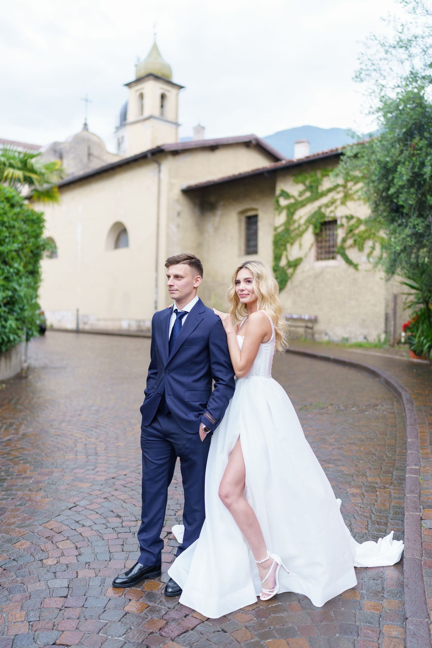 A couple poses for wedding photos outside a Mediterranean-style church with a bell tower and curved brick path.