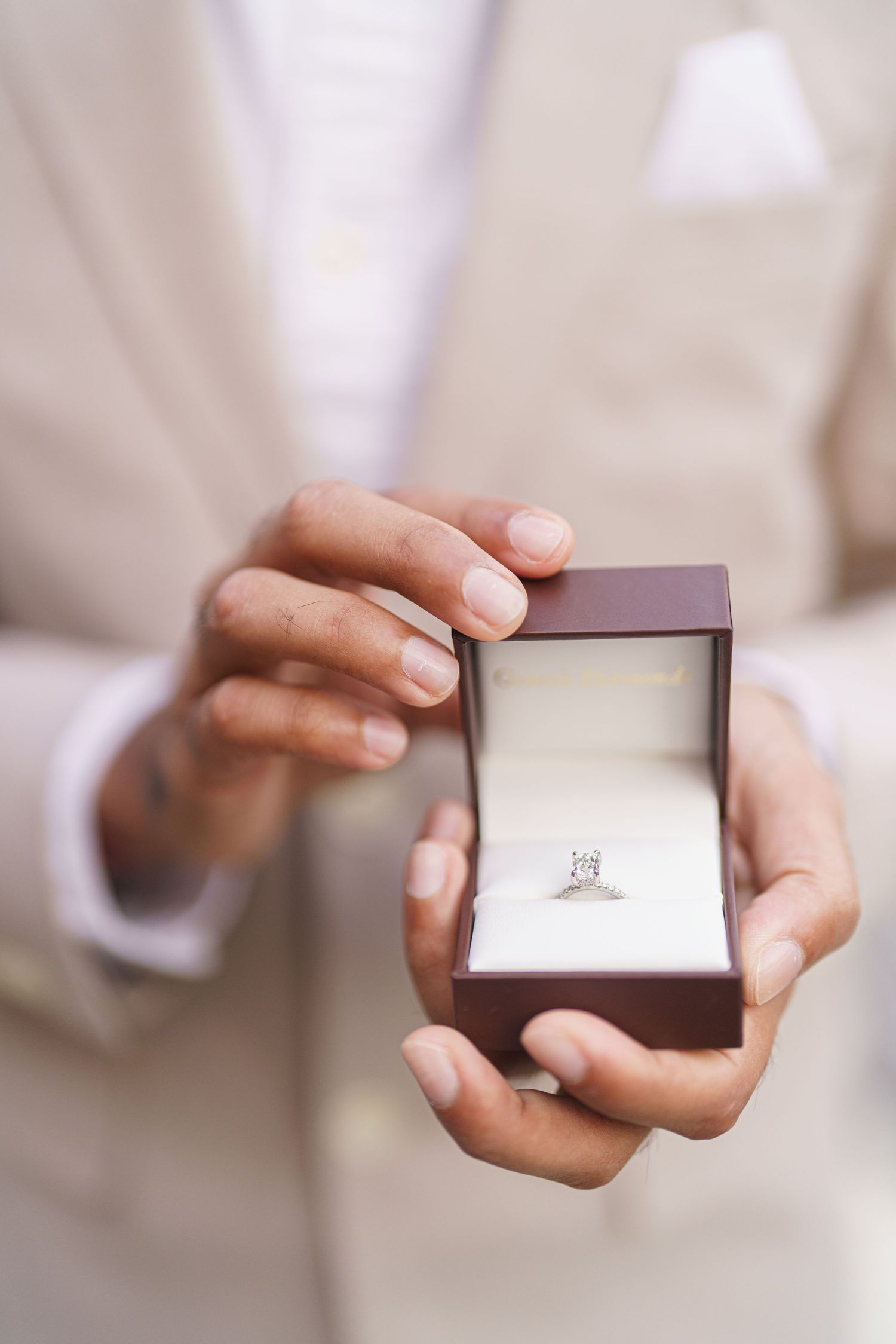 Hands hold a small brown jewelry box displaying a delicate diamond engagement ring against a soft white background.