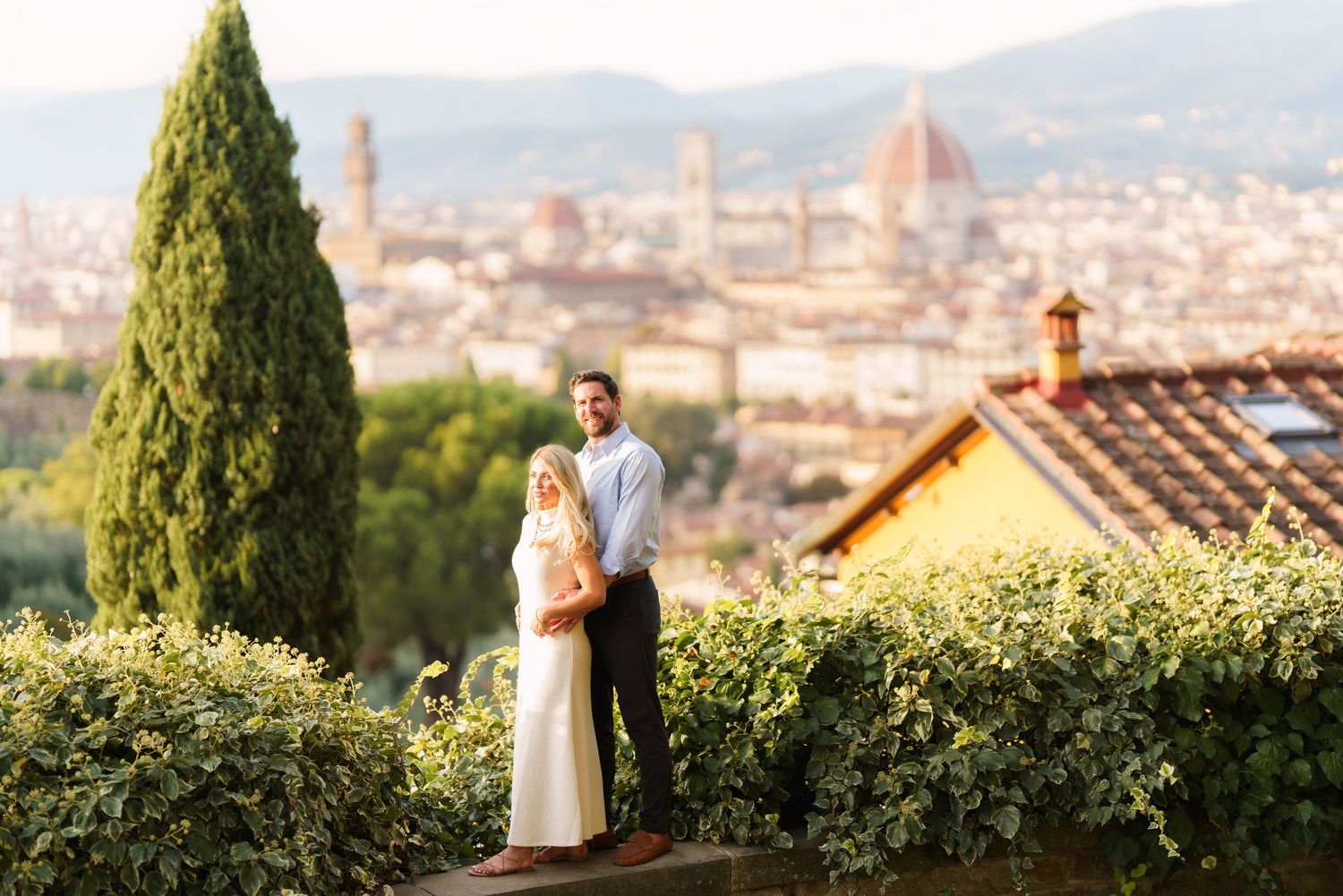 A couple embraces on a hilltop overlooking Florence, Italy with the Duomo cathedral visible in the distance.