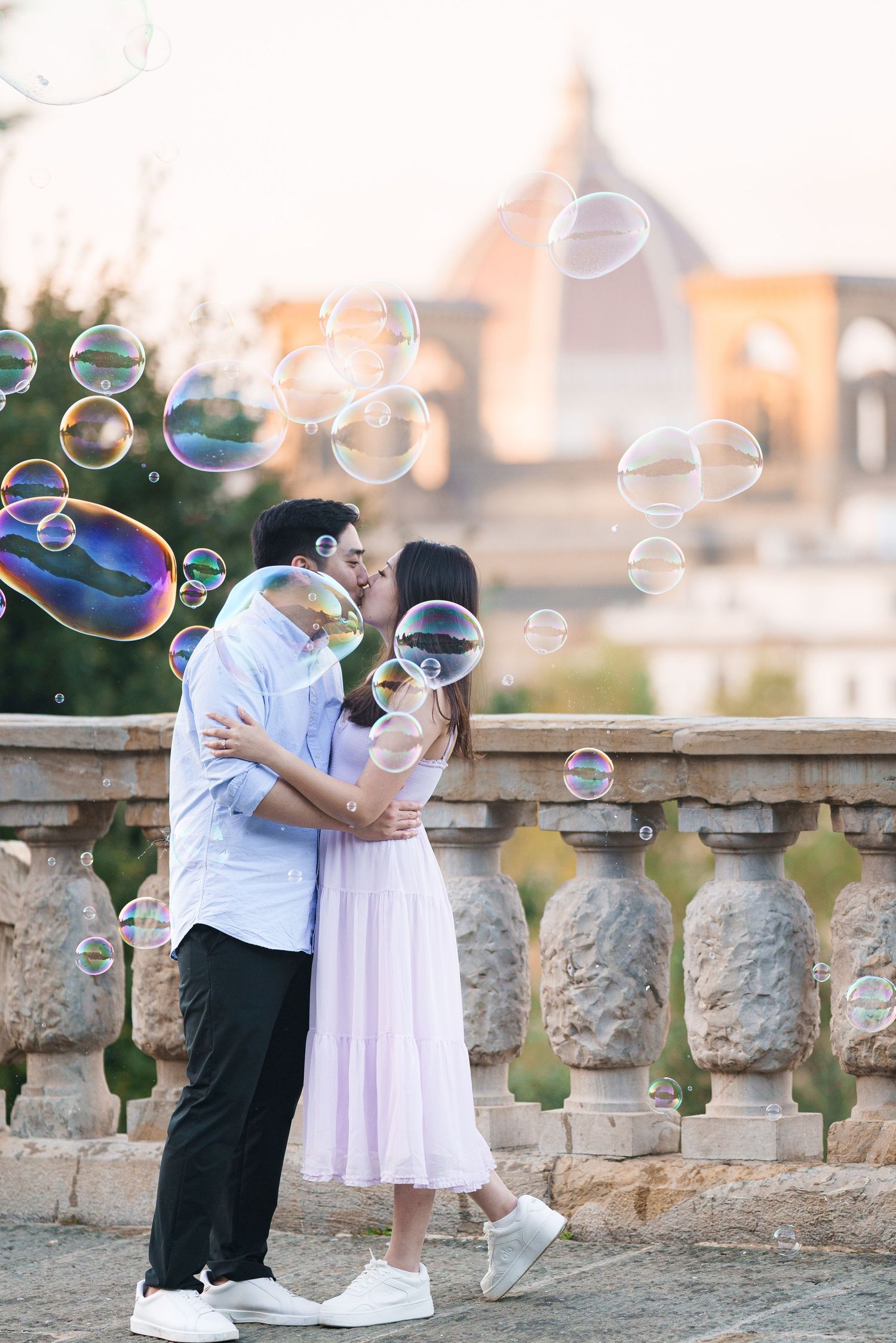 A romantic couple embraces on a stone balcony as iridescent soap bubbles float around them at sunset.