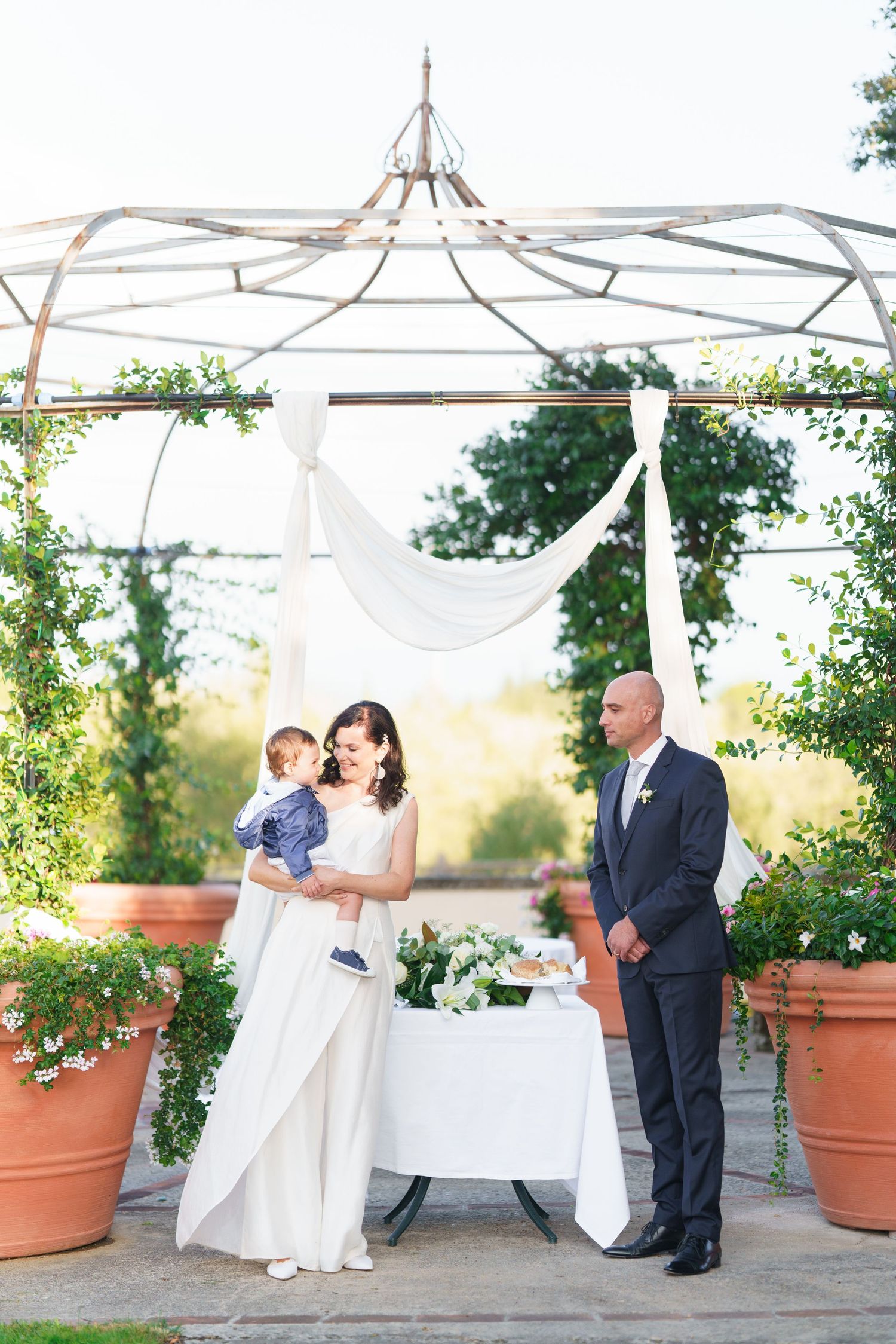 Elegant outdoor wedding scene with white draping and large terracotta planters on stone patio.