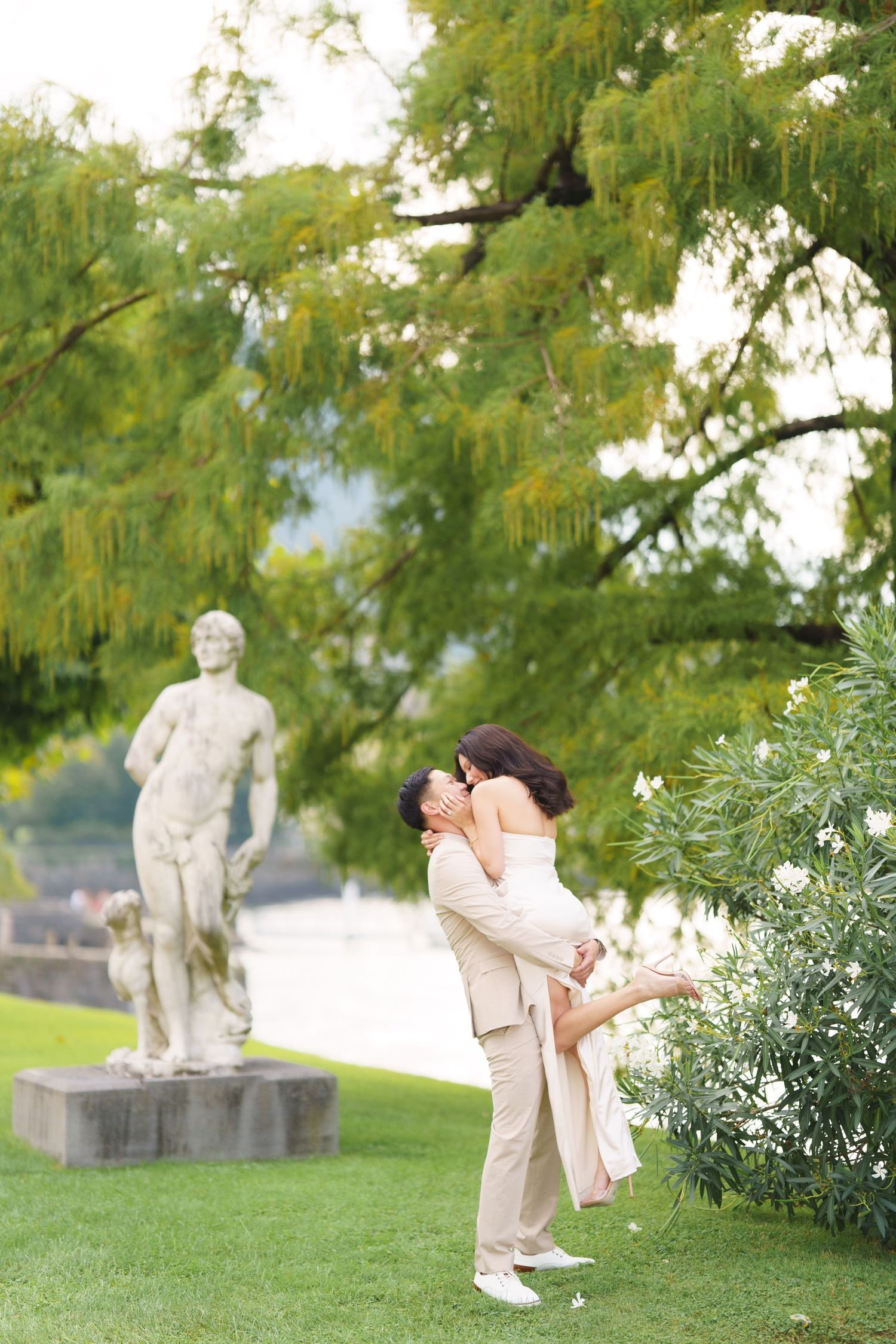 A romantic couple shares an intimate moment in a garden with a classical statue in the background.