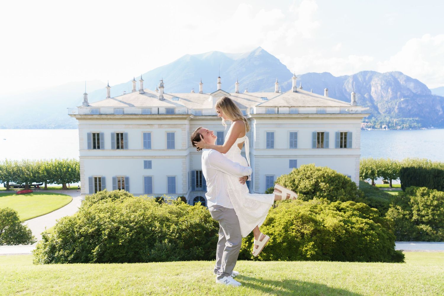 A romantic moment captured in front of a grand white villa with mountains and lake in the background.