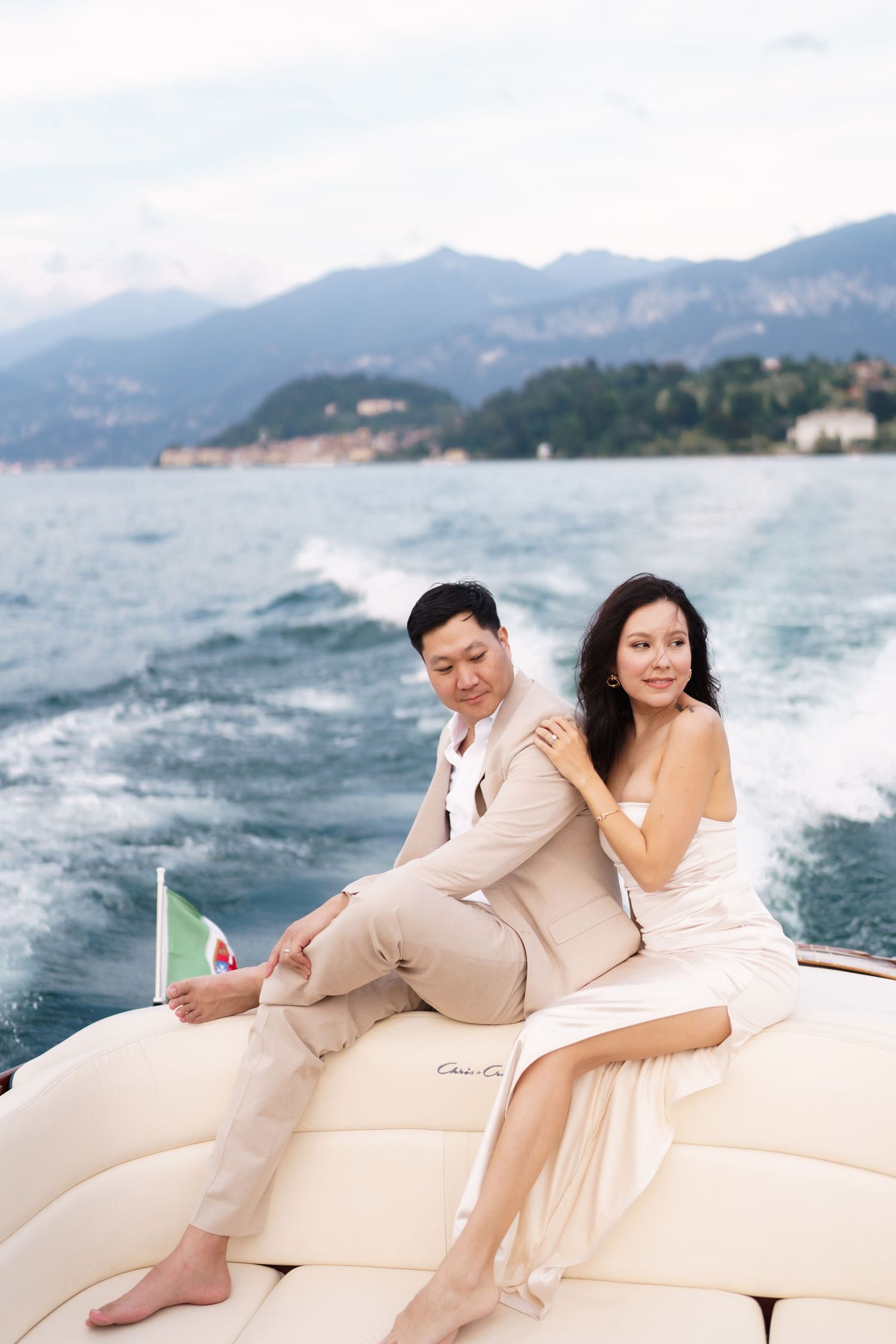 A couple relaxes on a luxury yacht with mountains and ocean in the background on a sunny day.
