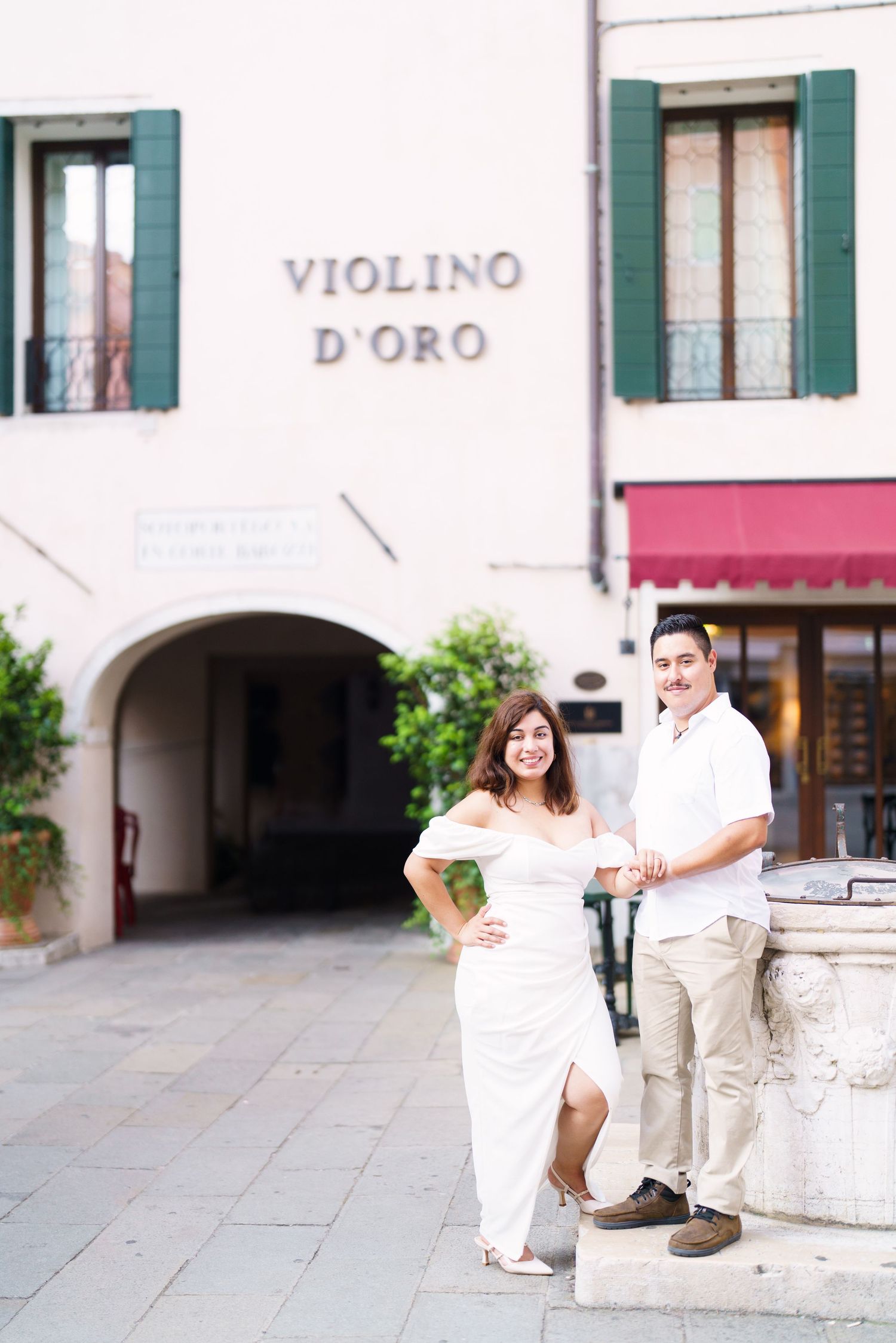 A couple in white outfits sits on stone steps in front of a charming Italian storefront with green shutters.