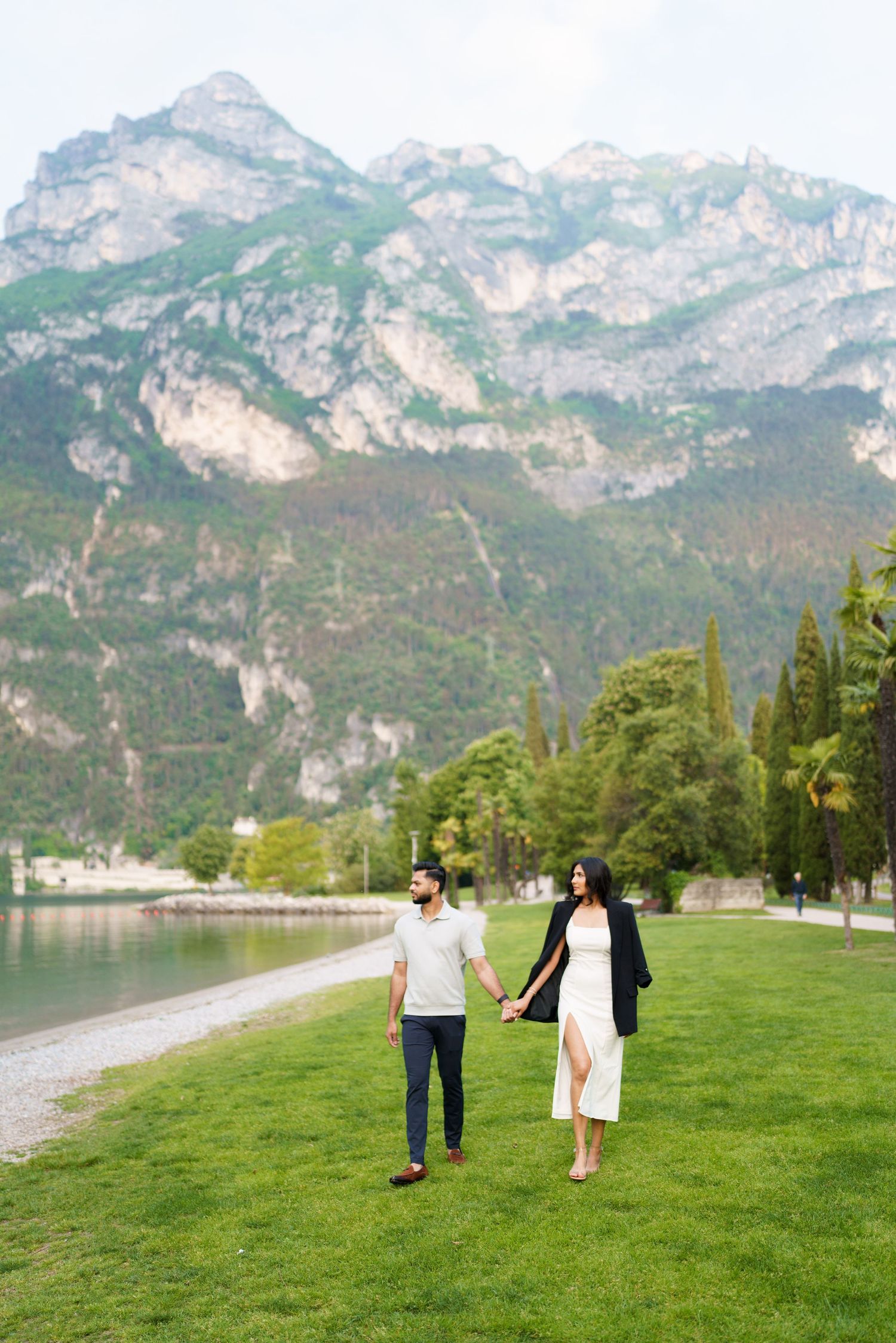 A couple walks hand in hand on green grass with majestic mountain peaks and a lake in the scenic backdrop.