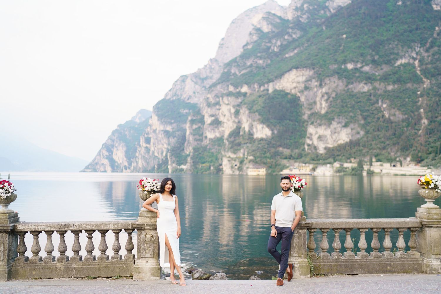 Couple poses on scenic balcony overlooking Lake Garda in Italy with mountains and blue water in background.