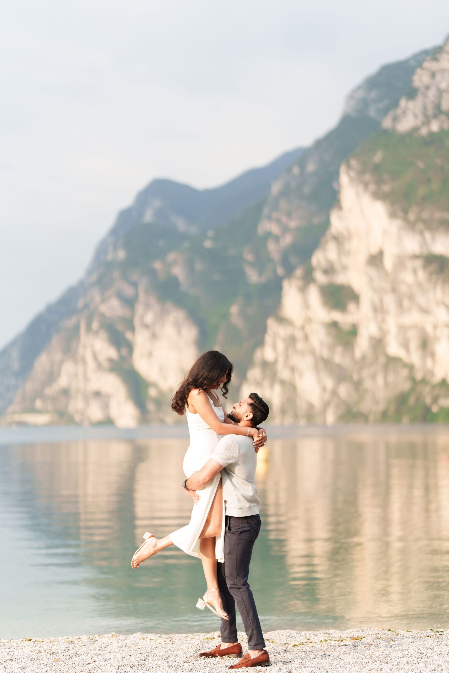 A couple embraces on a scenic lakeside beach with towering limestone cliffs and mountains in the background.