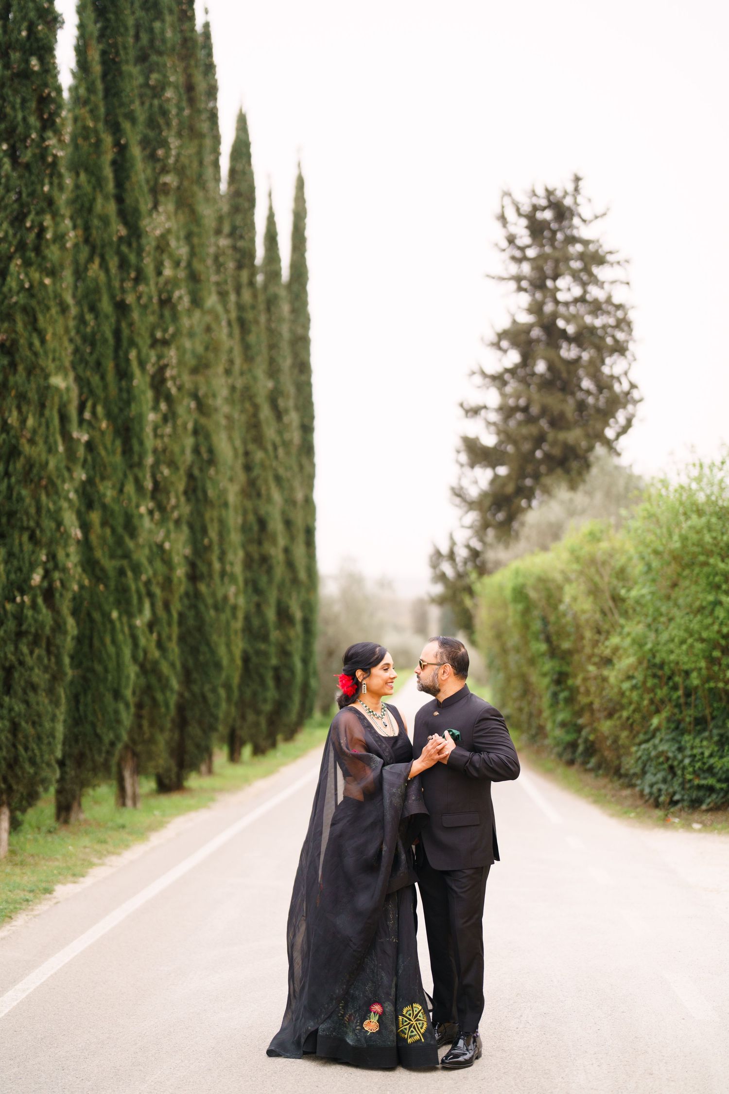 Romantic couple in black formal wear embracing on tree-lined pathway.