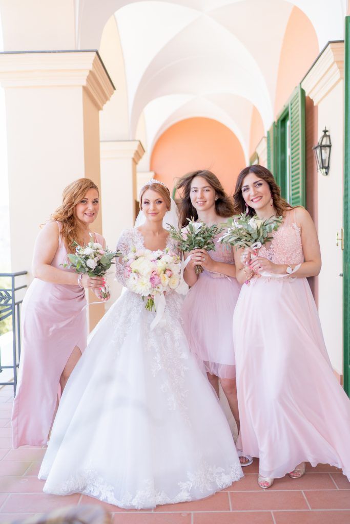 Wedding party in pink dresses and white gown gather for photos under arched colonnade.