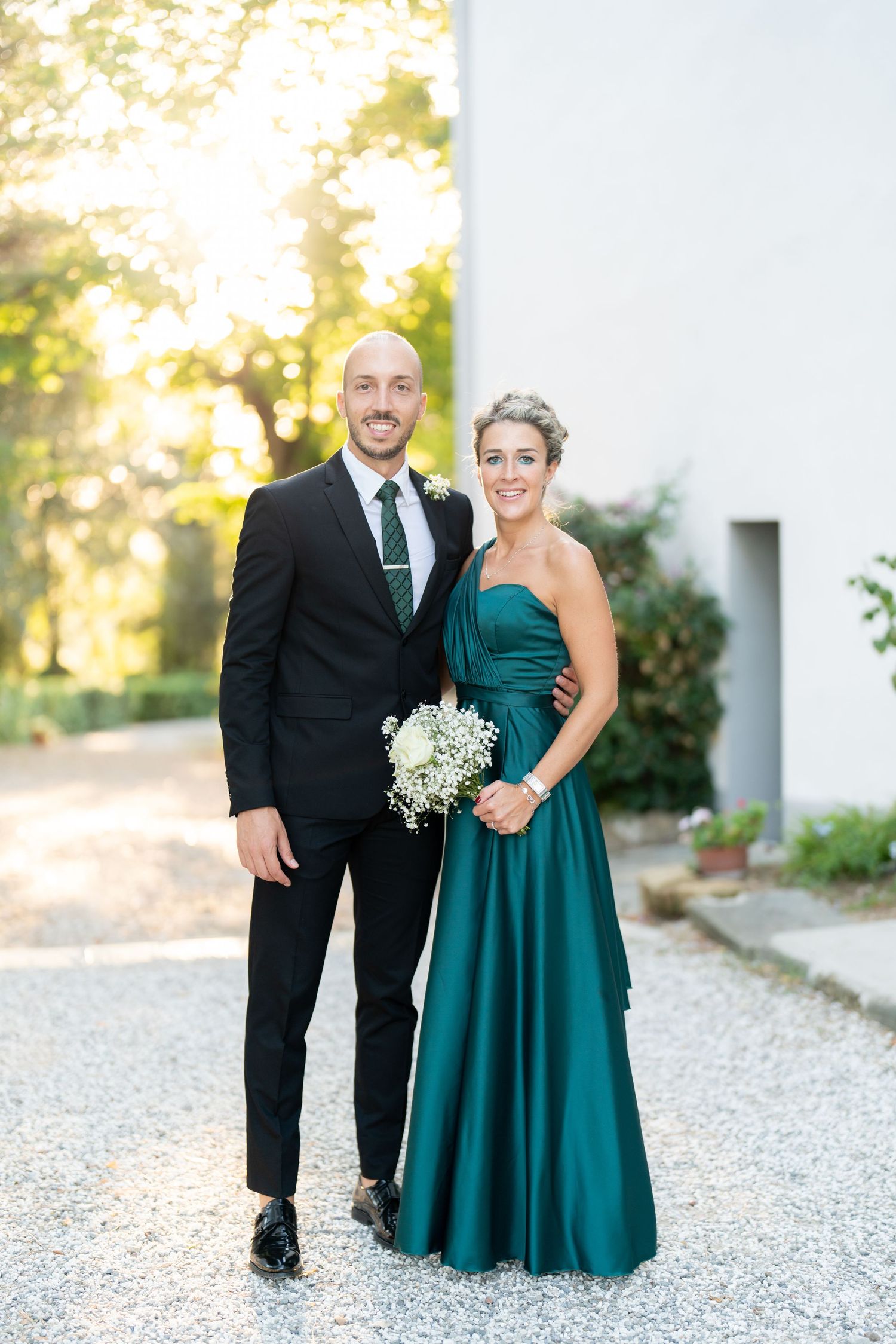 A formal couple poses outdoors in black tie and emerald dress with a bouquet.