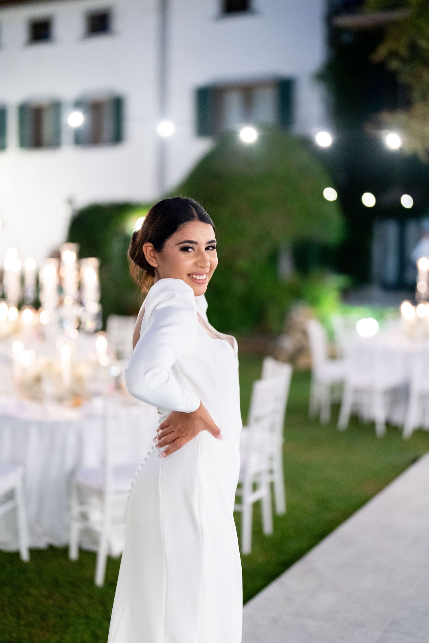 A white wedding dress is displayed against an elegant outdoor reception setting with string lights and tables.