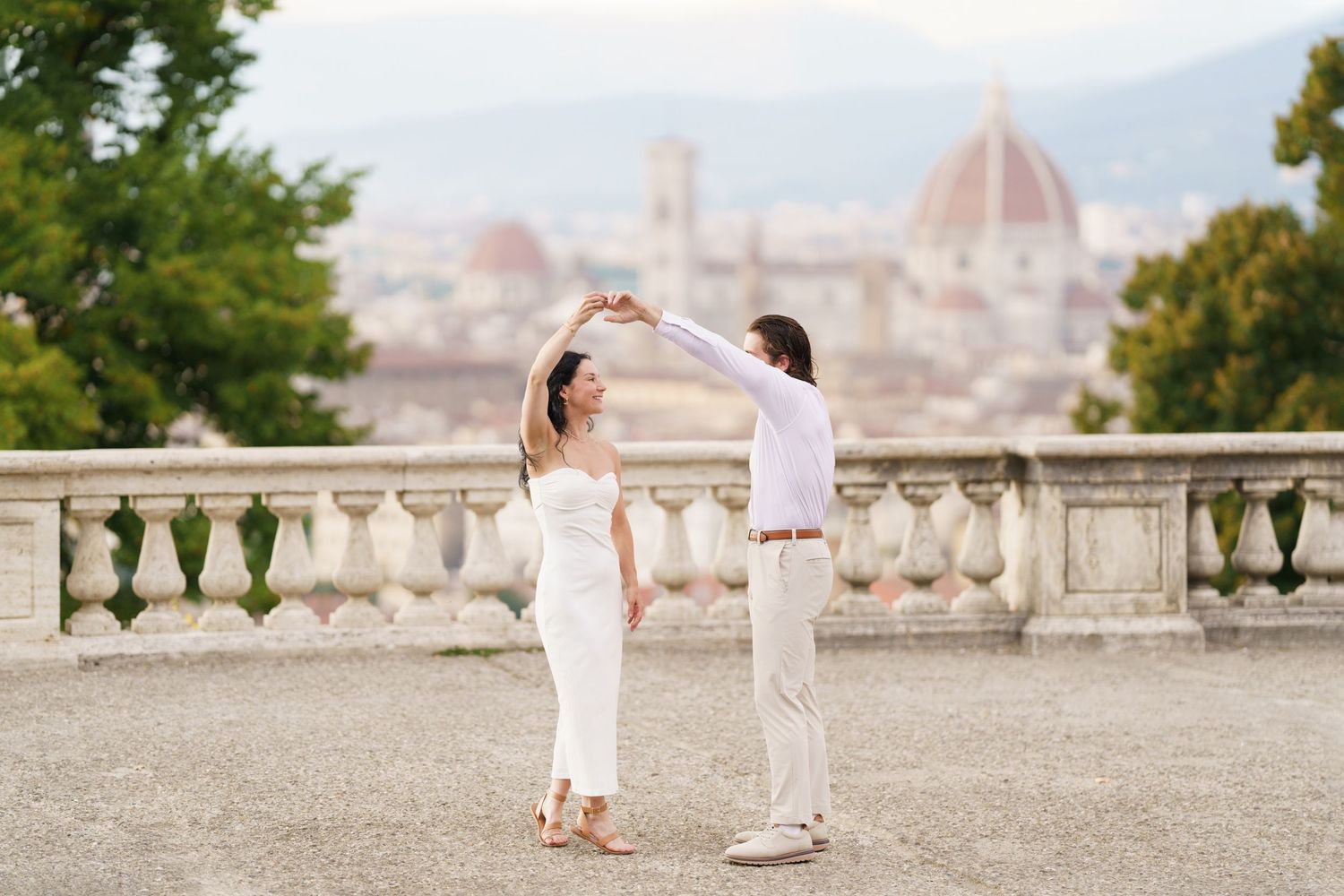 Two people in white outfits hold hands while walking along a scenic overlook with views of Florence, Italy in the background.