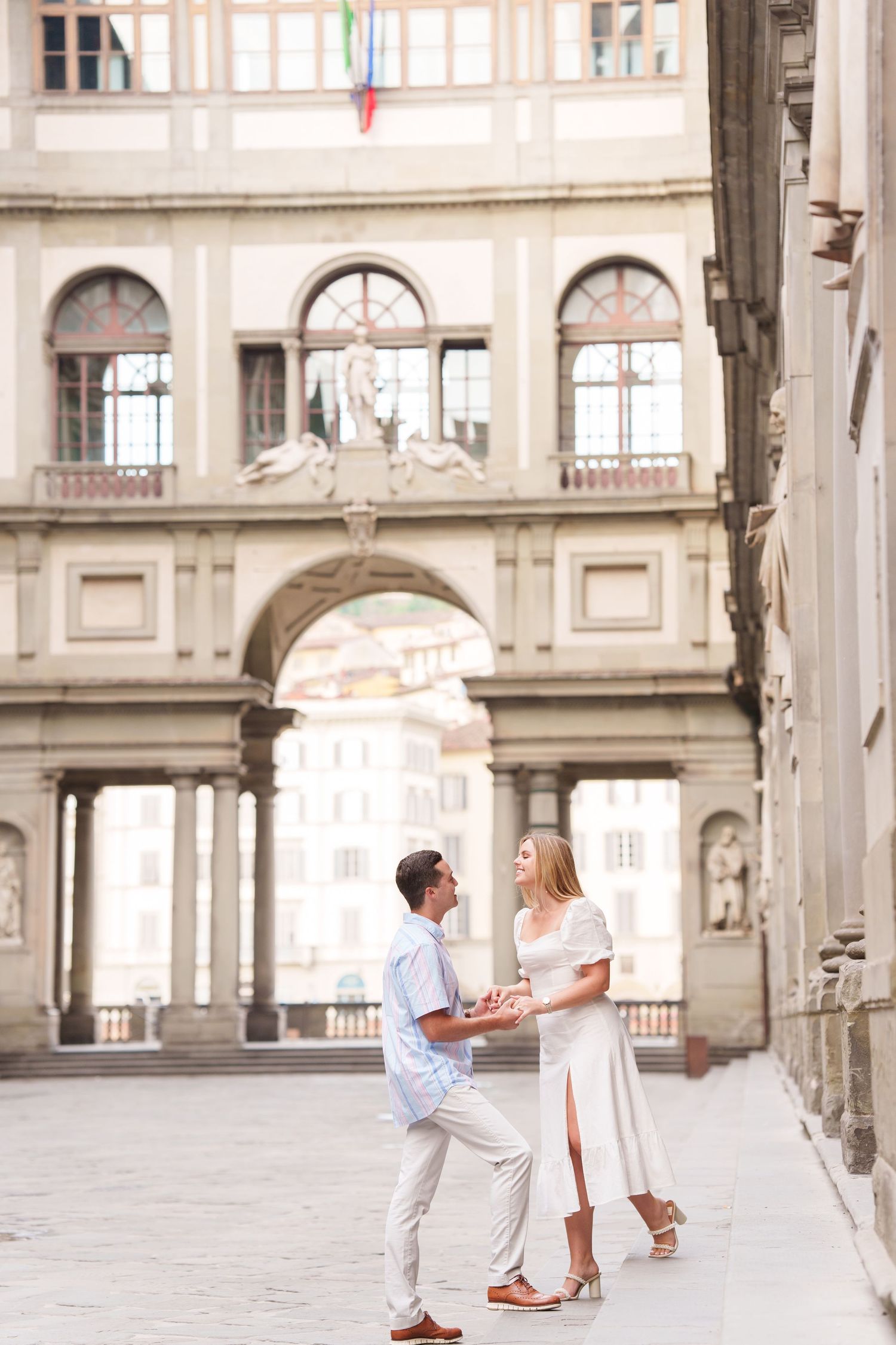 A couple in white outfits shares a romantic moment in a historic European courtyard with ornate architecture.