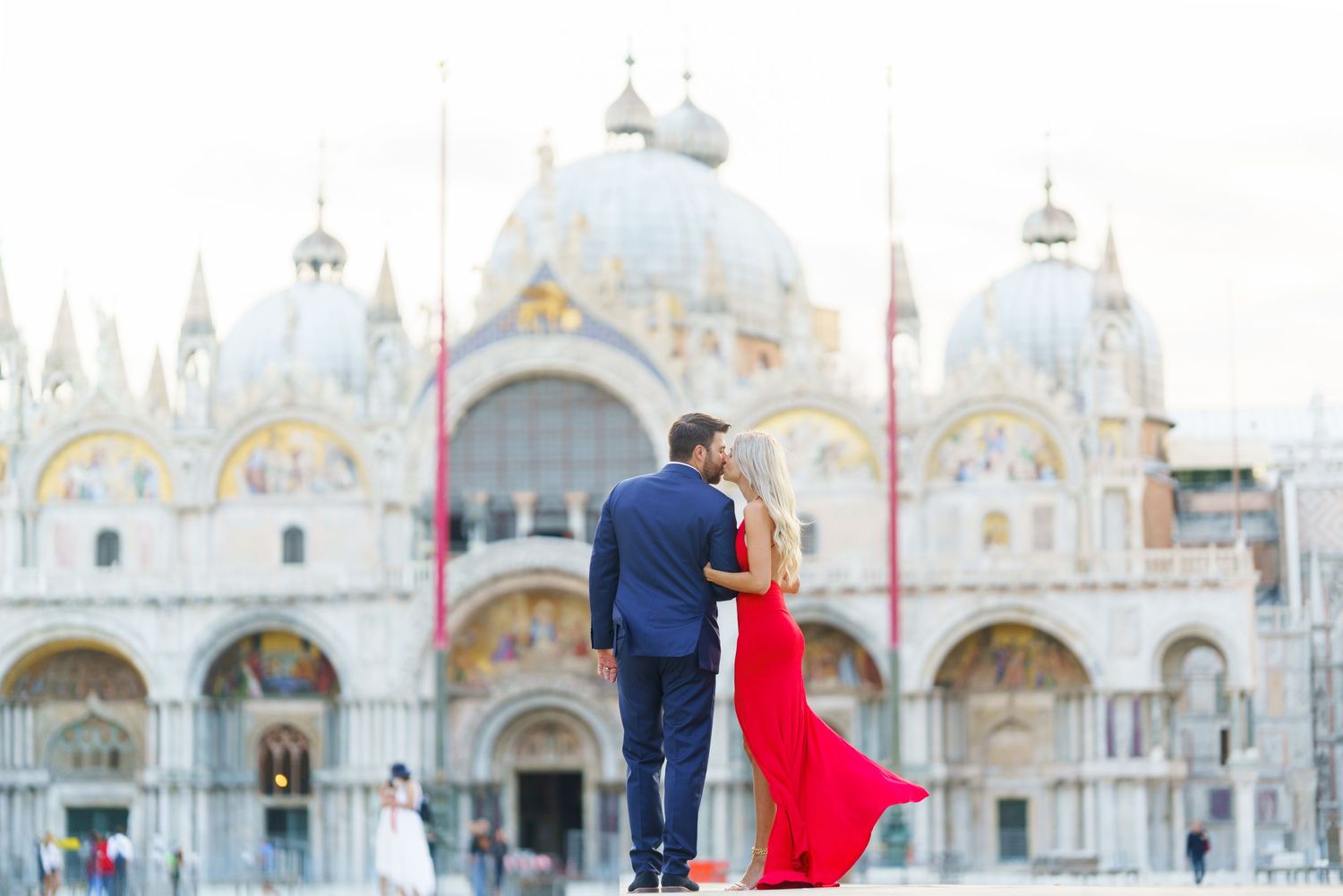 A couple in blue and red attire embrace while standing in front of St. Mark's Basilica in Venice, Italy.