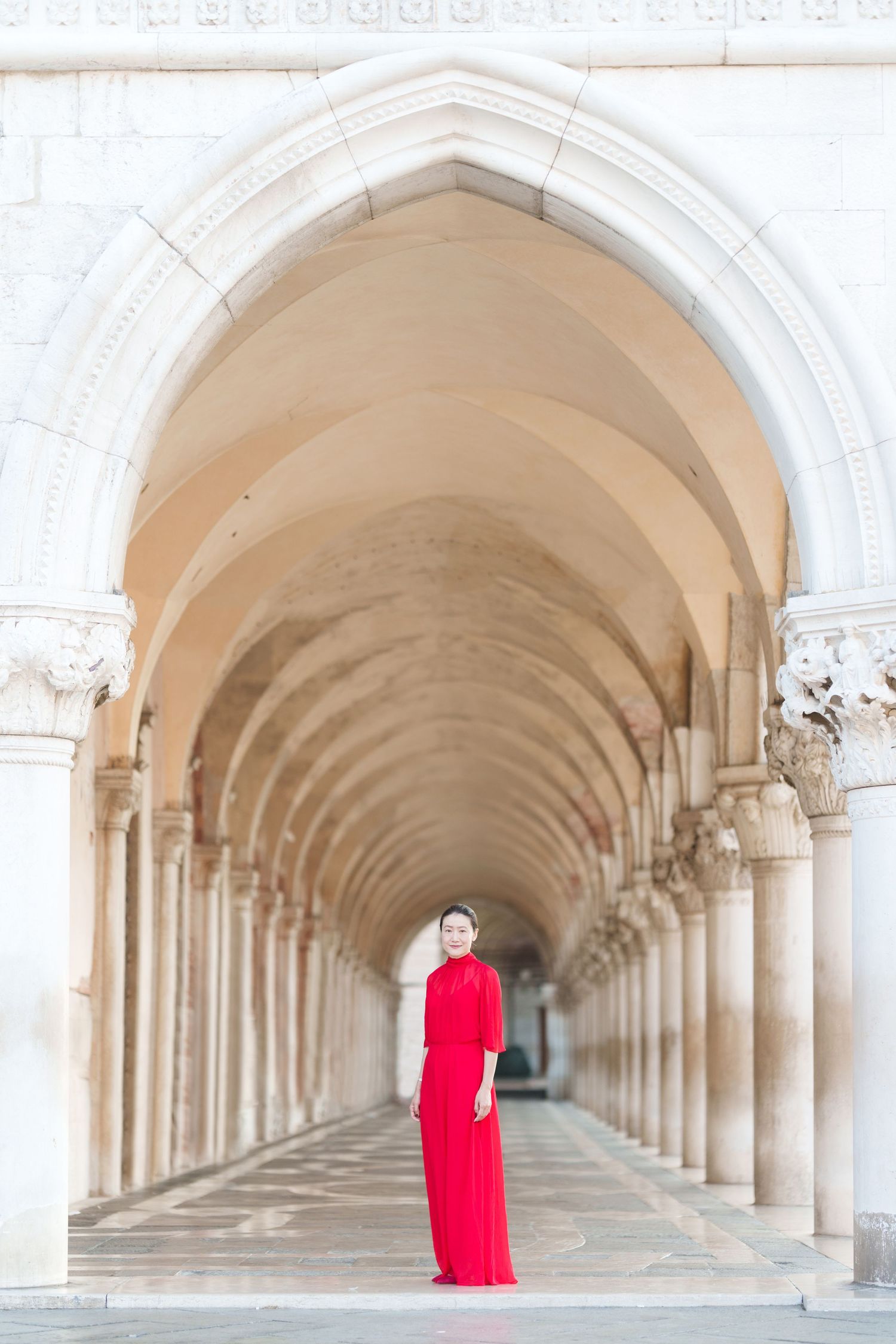 Woman in red gown standing under ornate arched colonnade in Venice, Italy.