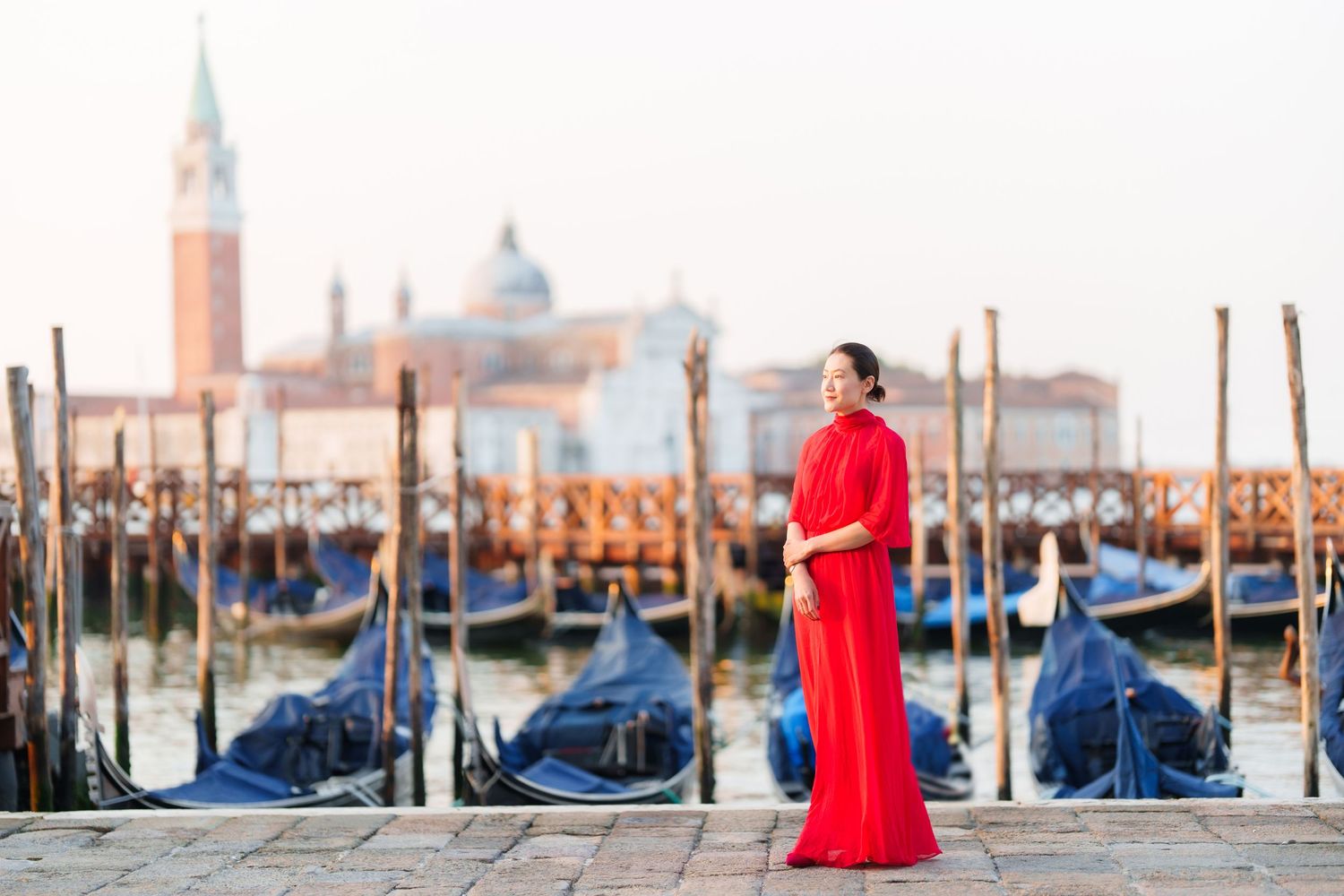 Woman in red dress standing by Venice gondolas with San Giorgio Maggiore church in background.