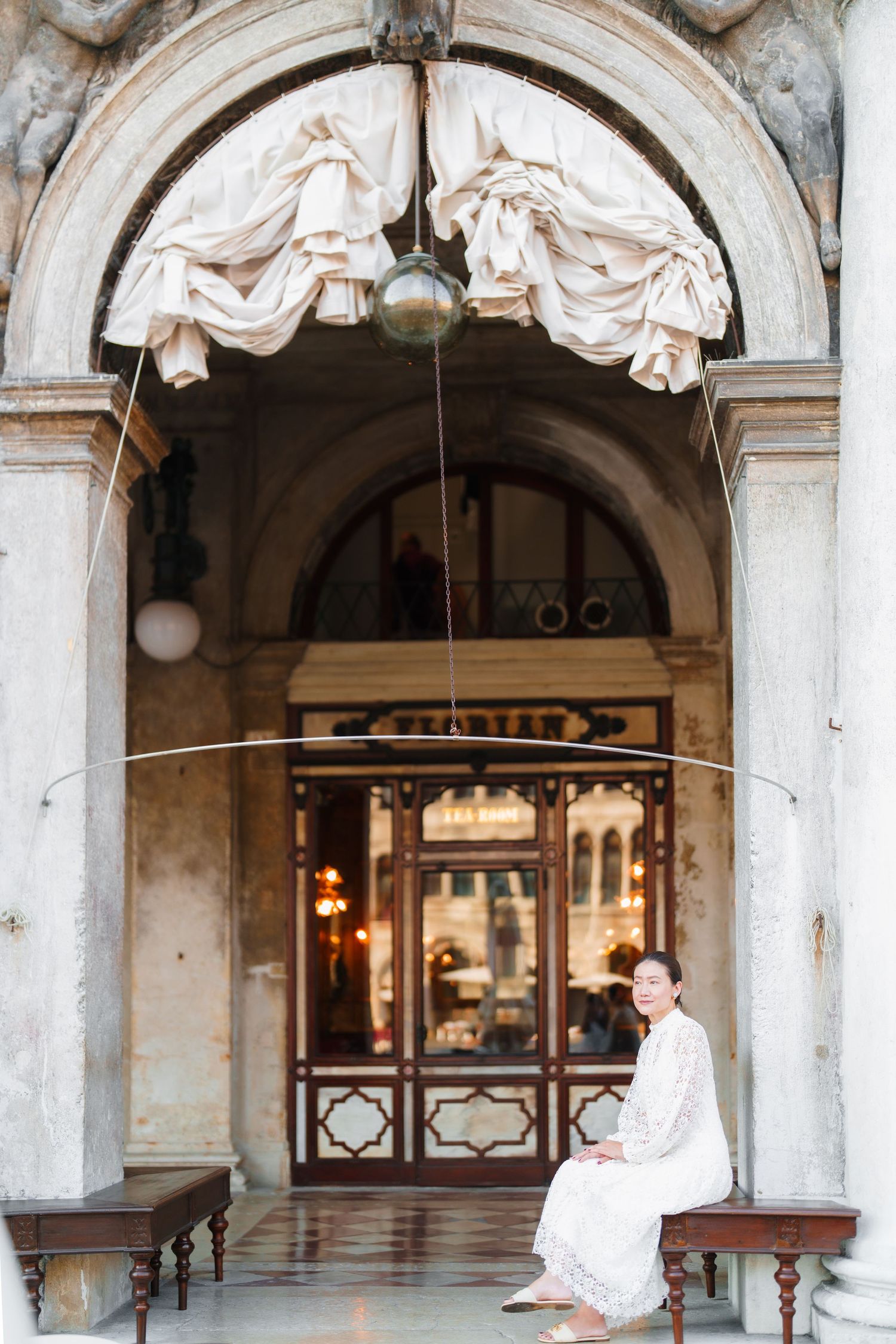 Woman in white dress sitting on bench under ornate arched entrance of historic Venetian building.