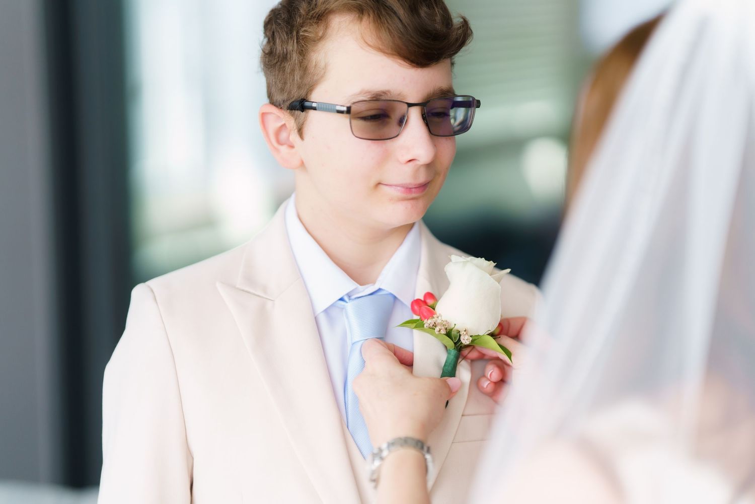 Groom in beige suit receiving boutonniere from bride in white veil at wedding ceremony.