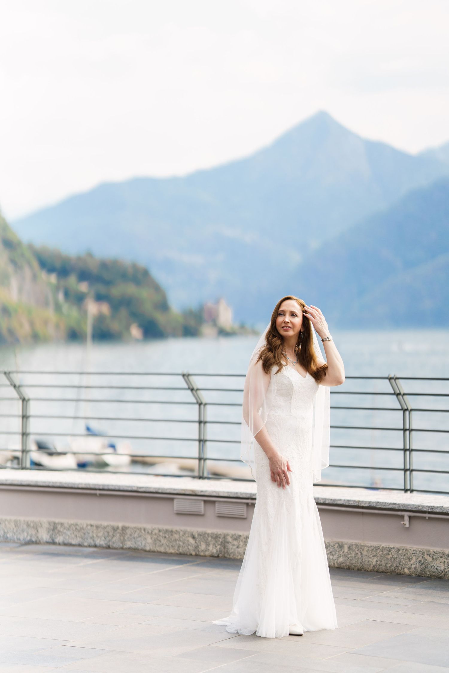 Bride in white gown poses on lakeside terrace with mountain backdrop.