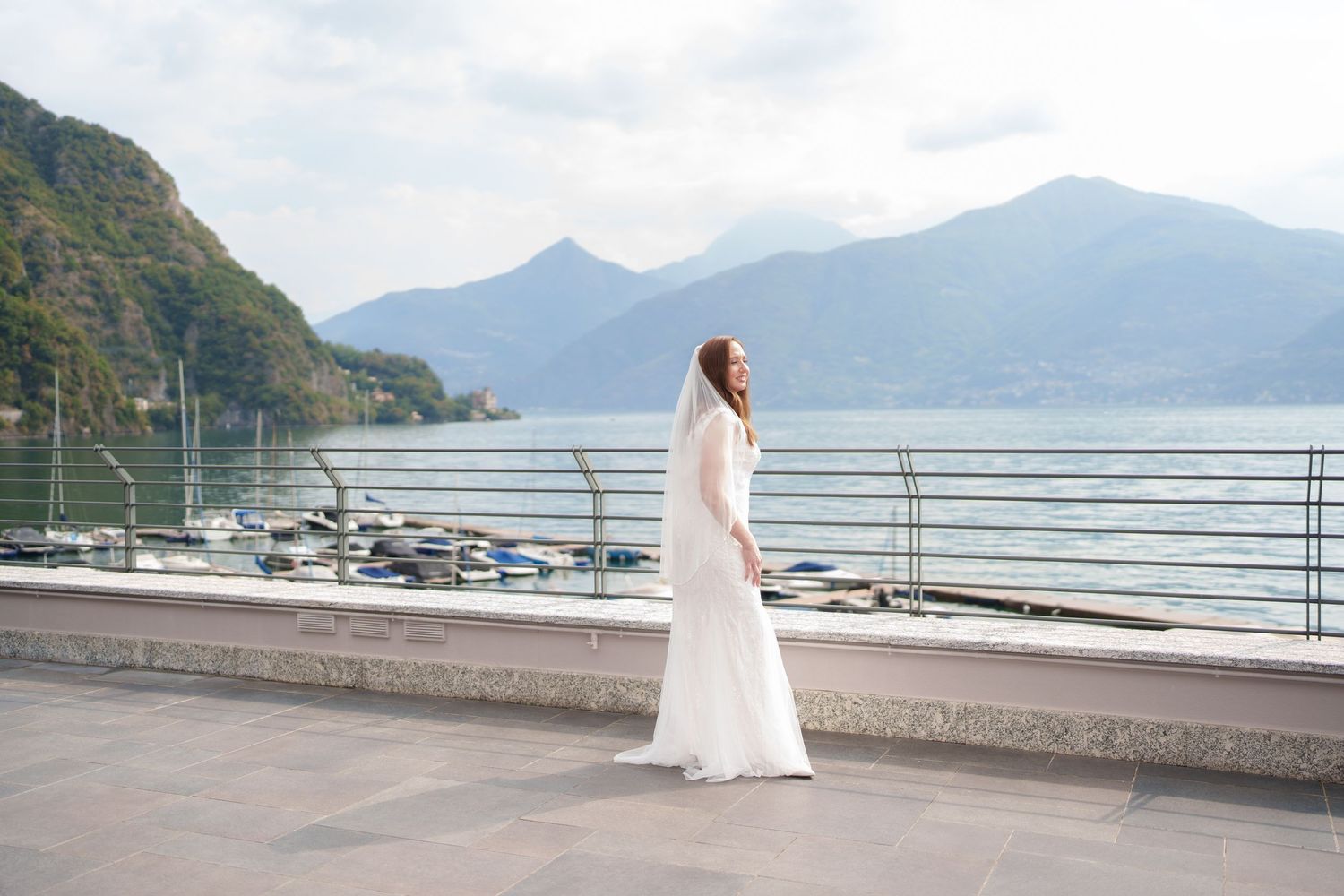 Bride in white gown stands on lakeside promenade with mountain backdrop at Lake Como, Italy.