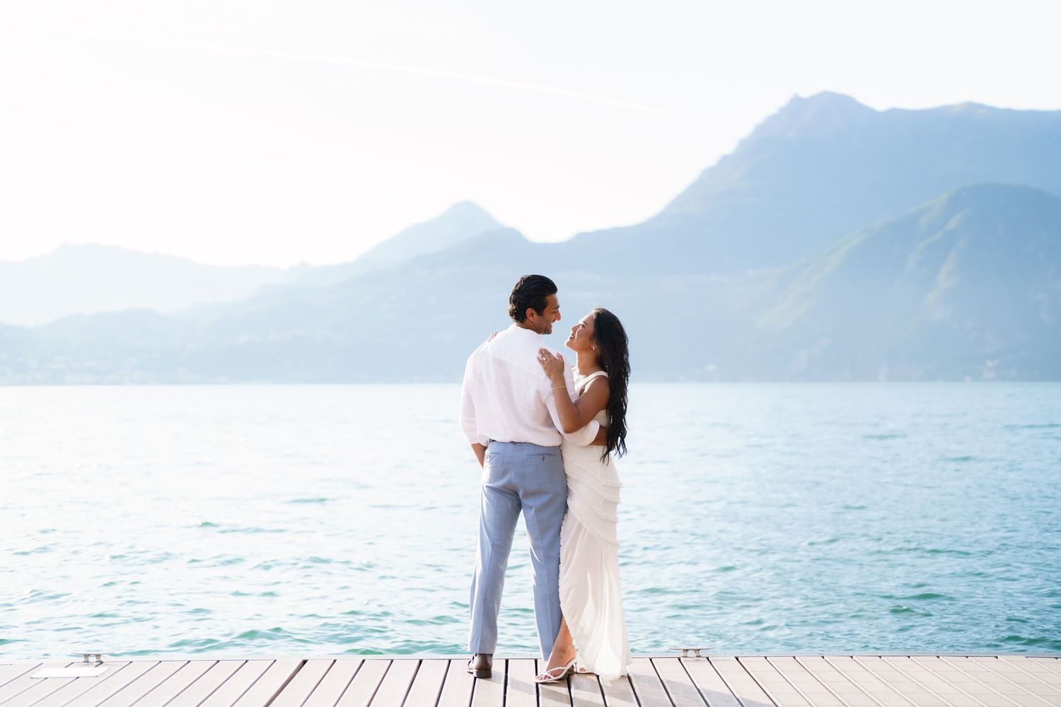 Couple in white attire sharing a romantic moment on a wooden dock overlooking a serene mountain lake.