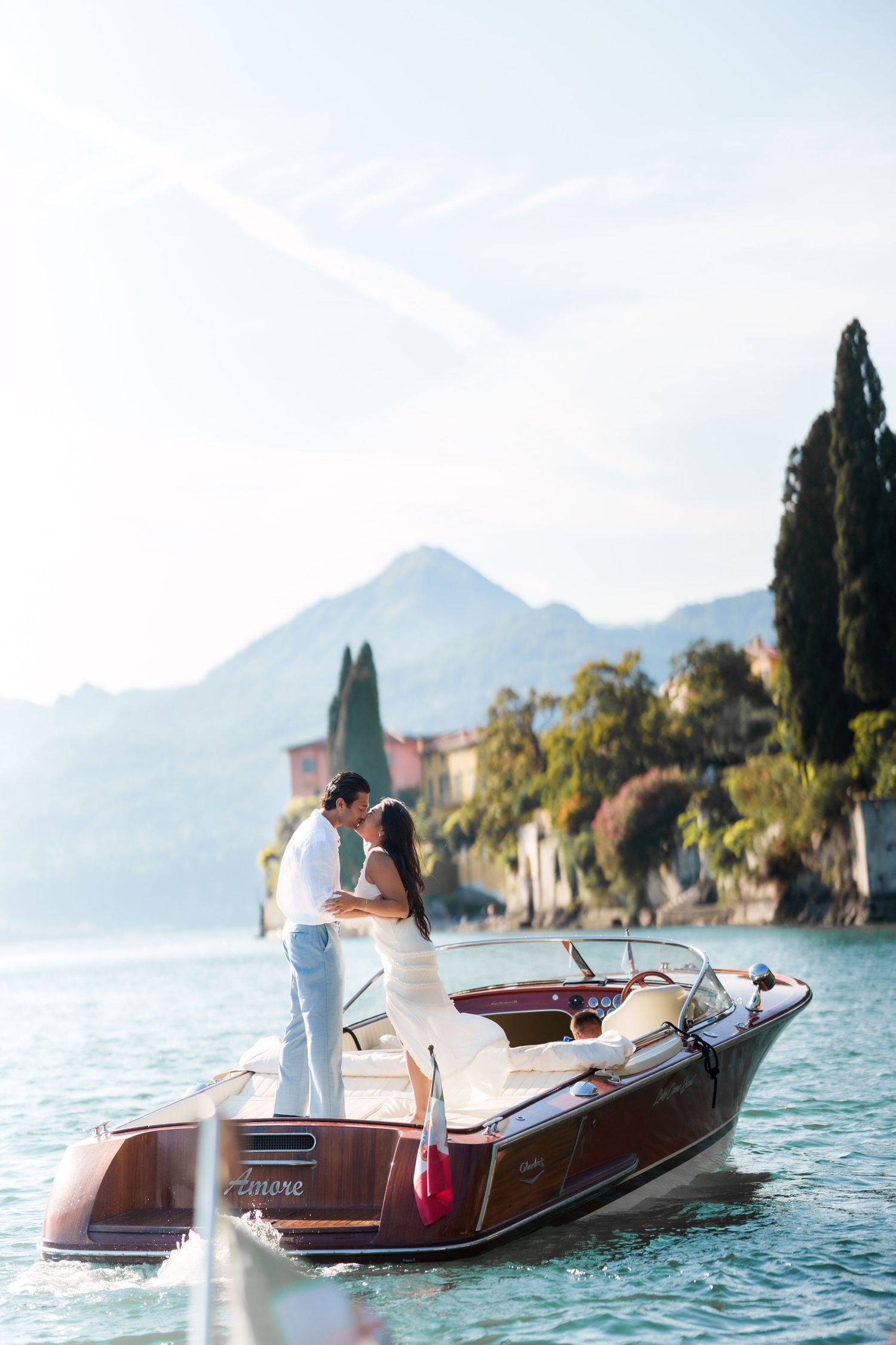 Couple kissing on a wooden boat named Amore on Lake Como, Italy, with mountains and cypress trees in the background.