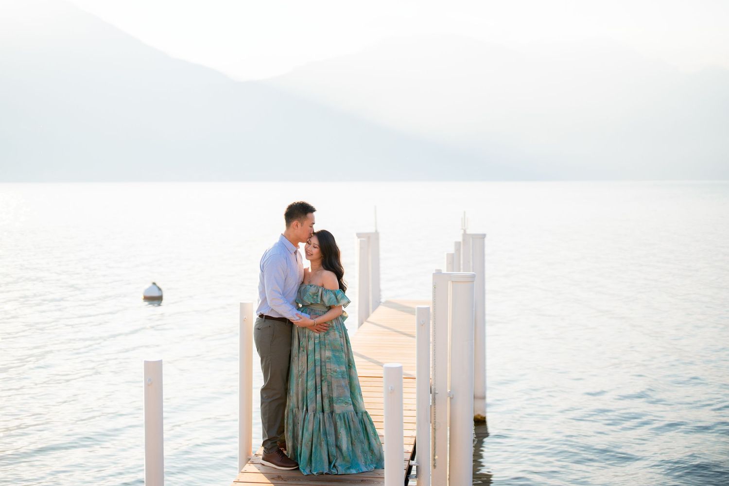 Couple embracing on a wooden dock overlooking calm lake waters with misty mountains in the background.