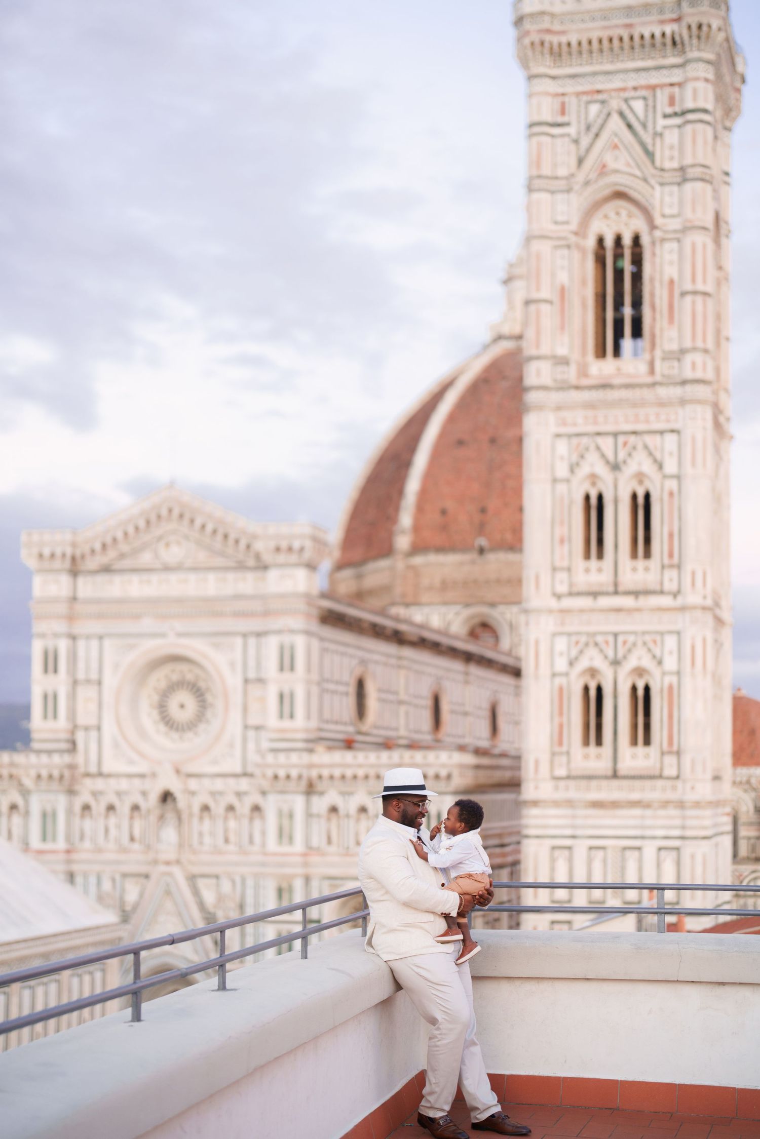 Man in white suit holding baby on rooftop terrace with Florence Cathedral dome and Giotto's Campanile backdrop.