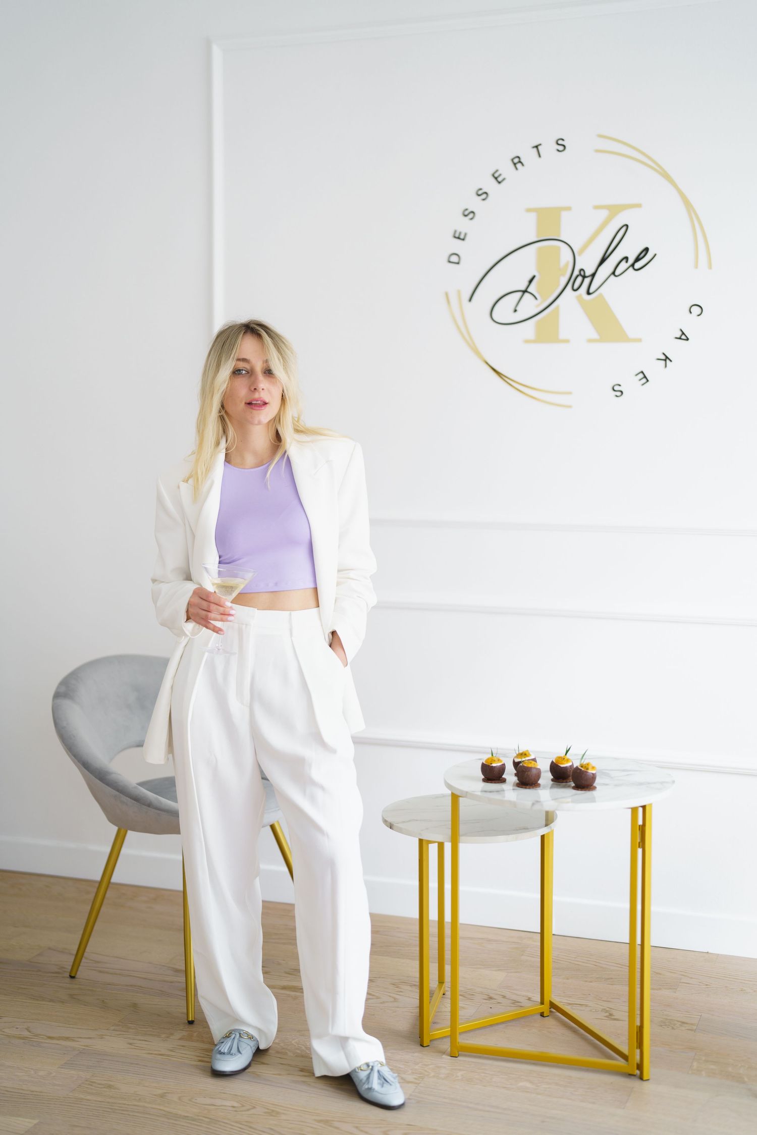 Blonde woman in white suit poses near gold table with cupcakes beside Dolce K Desserts Cakes logo on wall.