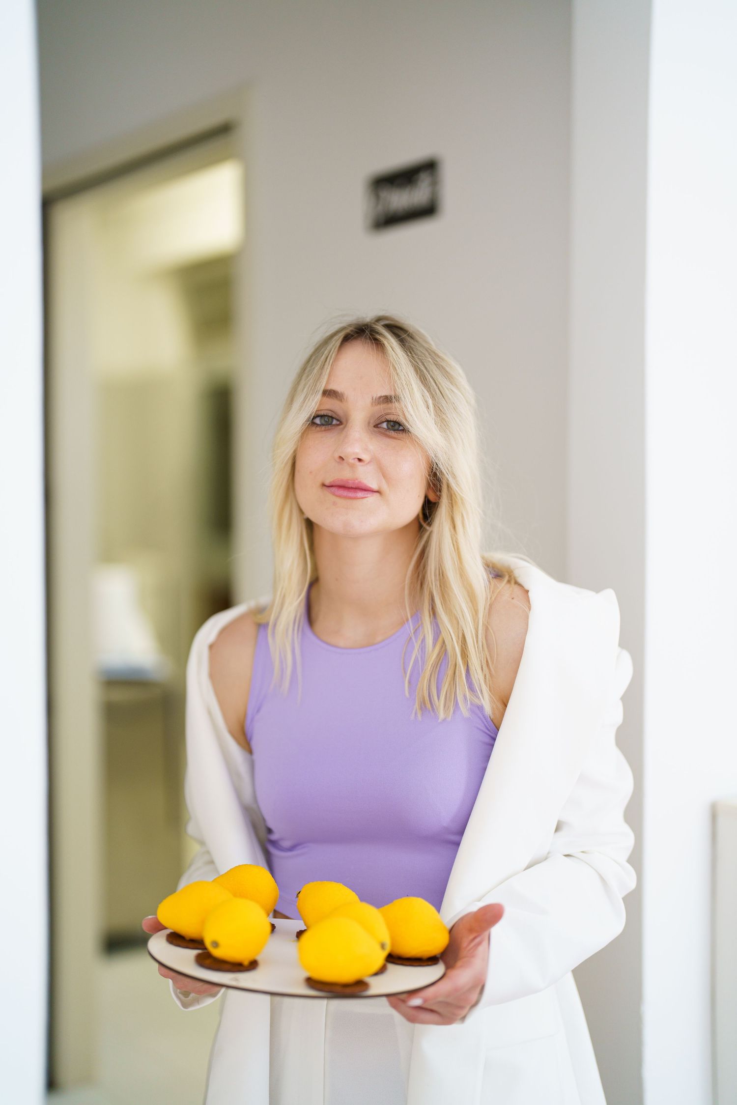 Blonde woman in purple top and white blazer holding a wooden plate with fresh yellow lemons indoors.