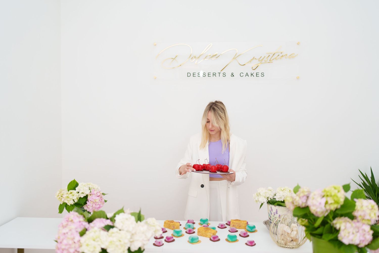 A woman in a white blazer arranges colorful macarons at a desserts and cakes shop with floral decor.